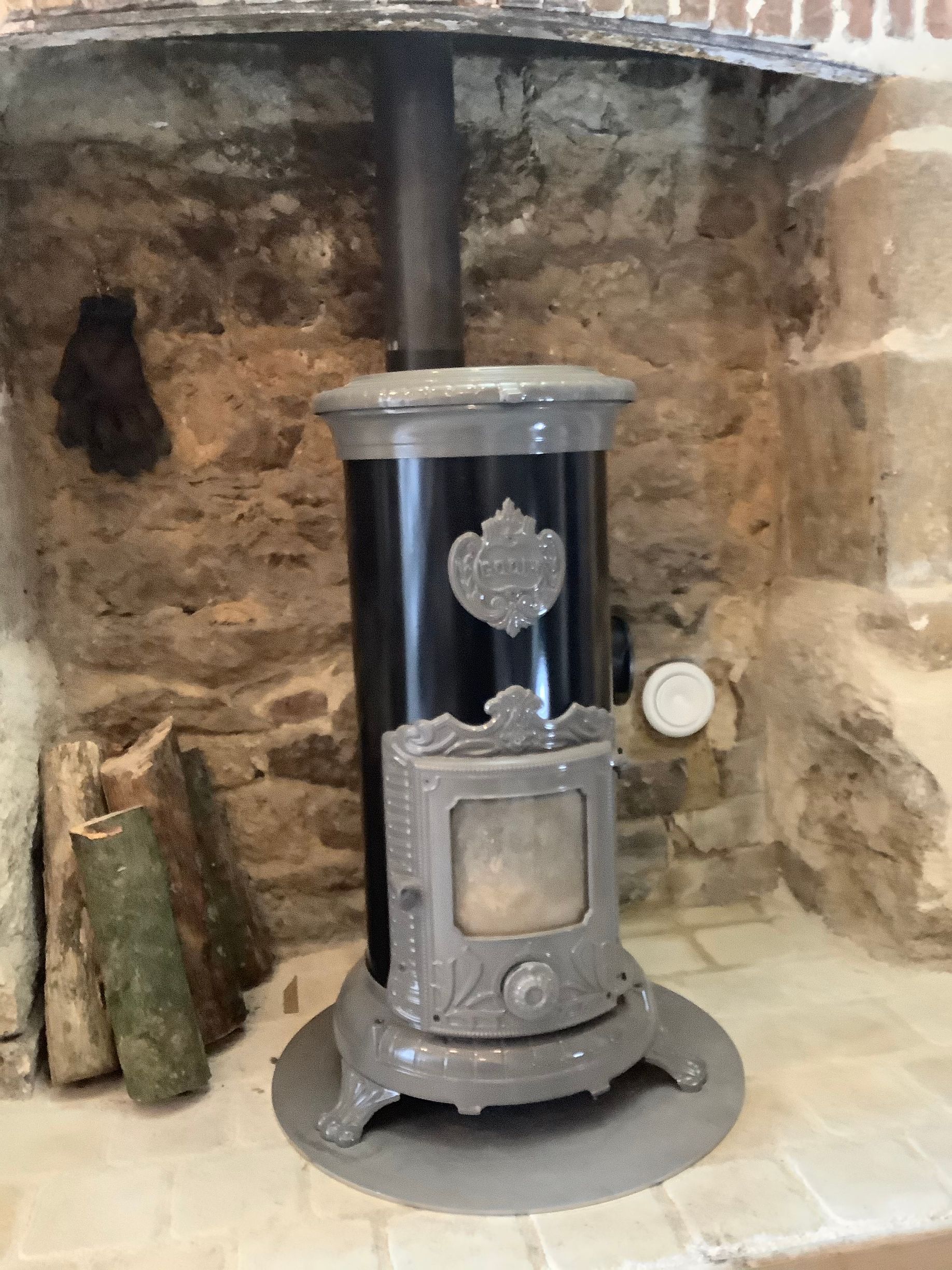 Un poêle à bois de style vintage, noir et gris, trône sur un foyer en pierre dans une cheminée rustique en briques.