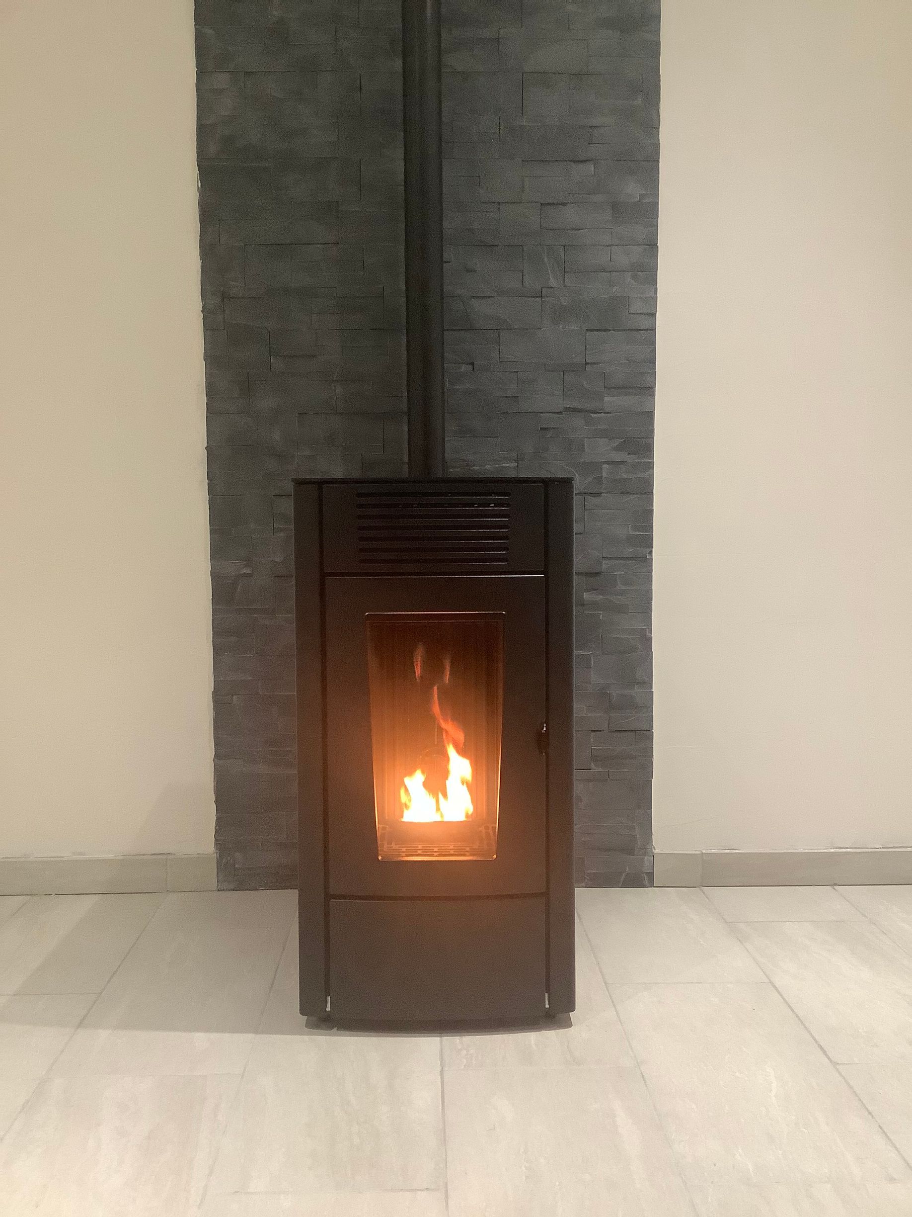 Un poêle à granulés noirs, dont la flamme est visible, est placé contre un mur en pierre grise sur un sol carrelé clair.
