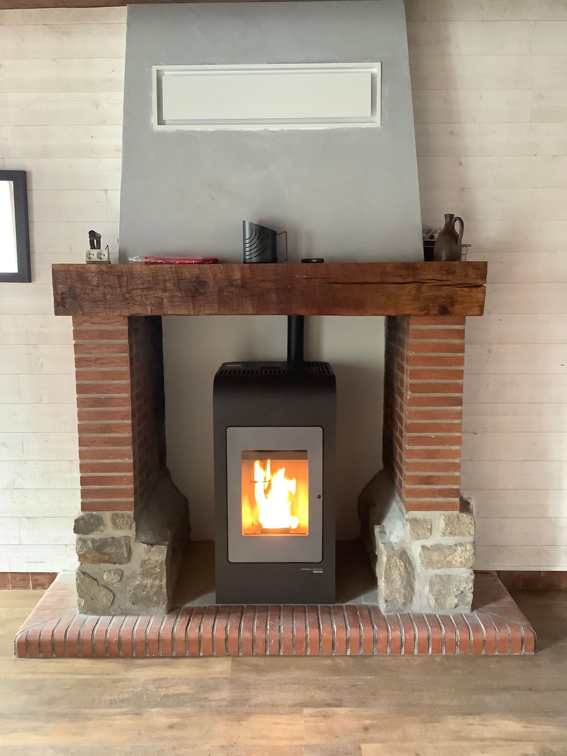 Un poêle à granulés à flamme visible est installé à l'intérieur d'une cheminée rustique en briques et en pierres, dotée d'un épais manteau de cheminée en bois.