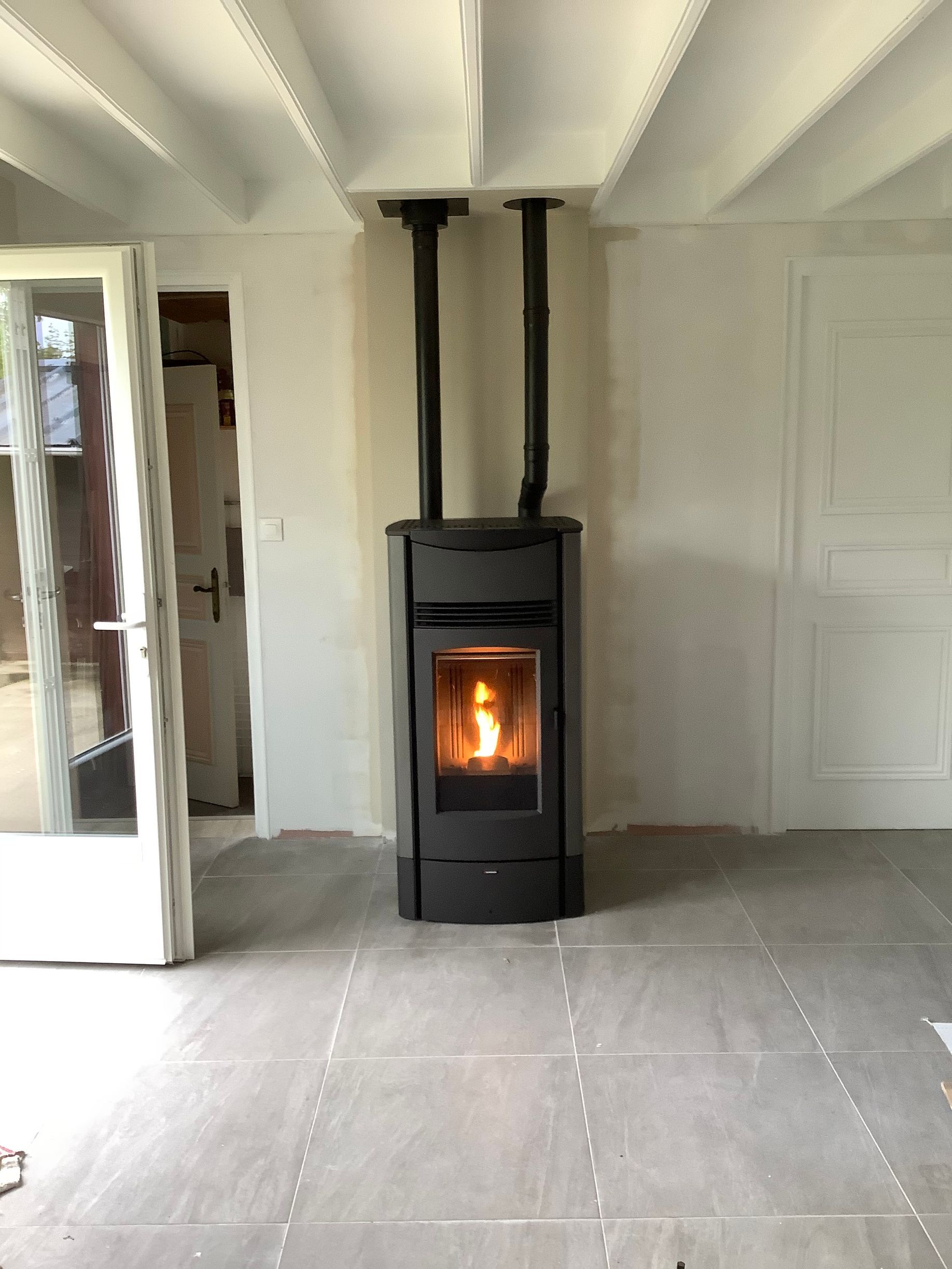 Un poêle à granulés noirs, dont la flamme est visible, est adossé à un mur blanc, deux tuyaux d'échappement noirs atteignant le plafond.