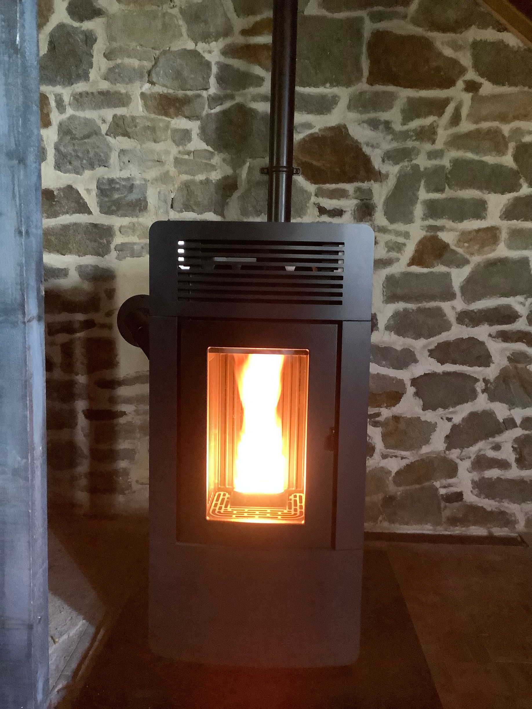 Un poêle à granulés en métal sombre, avec une flamme vive et visible à l'intérieur, est placé contre un mur de pierre rustique.