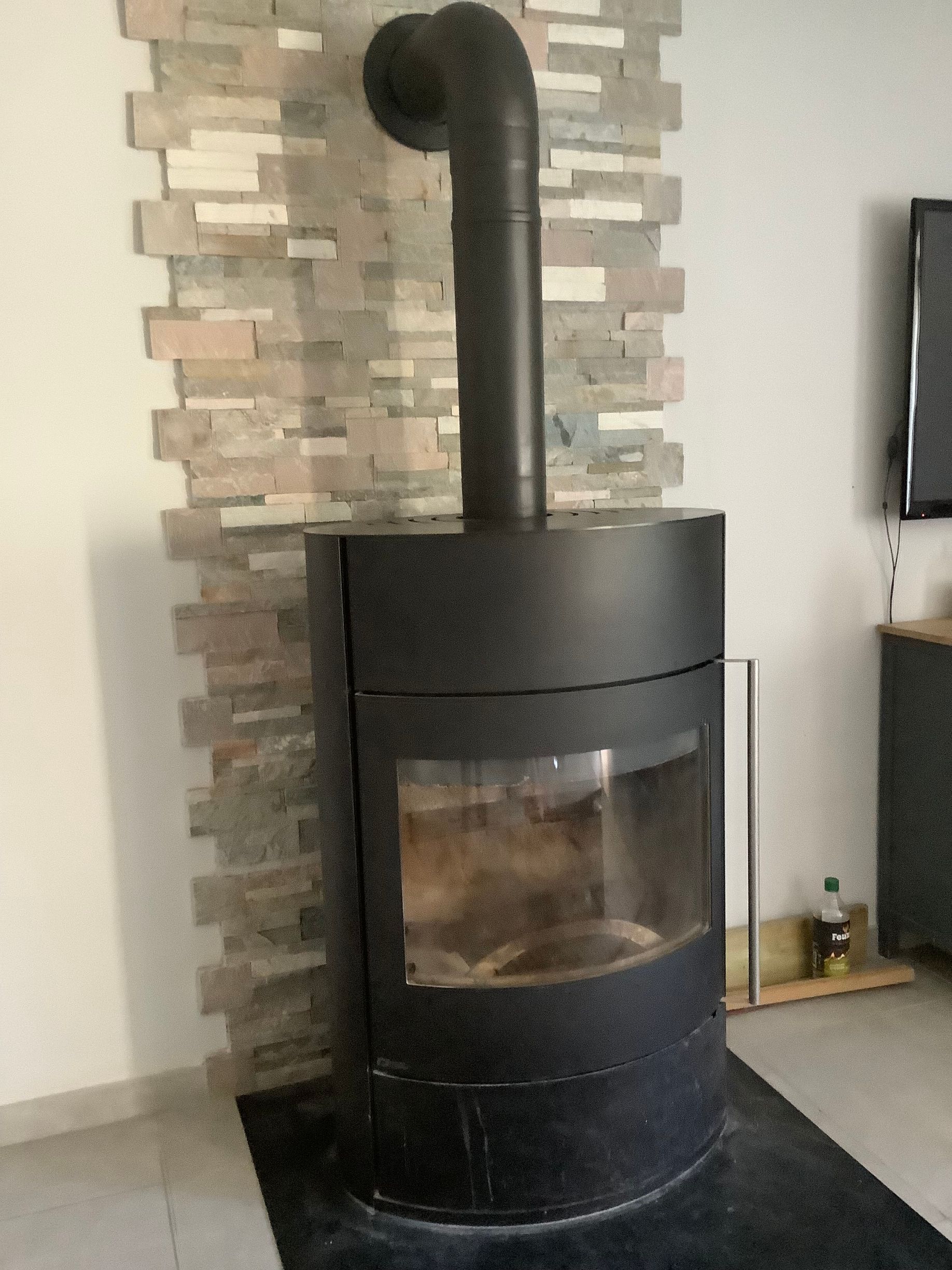 Un poêle à bois moderne noir est placé contre un mur d'accent en pierre beige à textures variées dans un espace de vie.