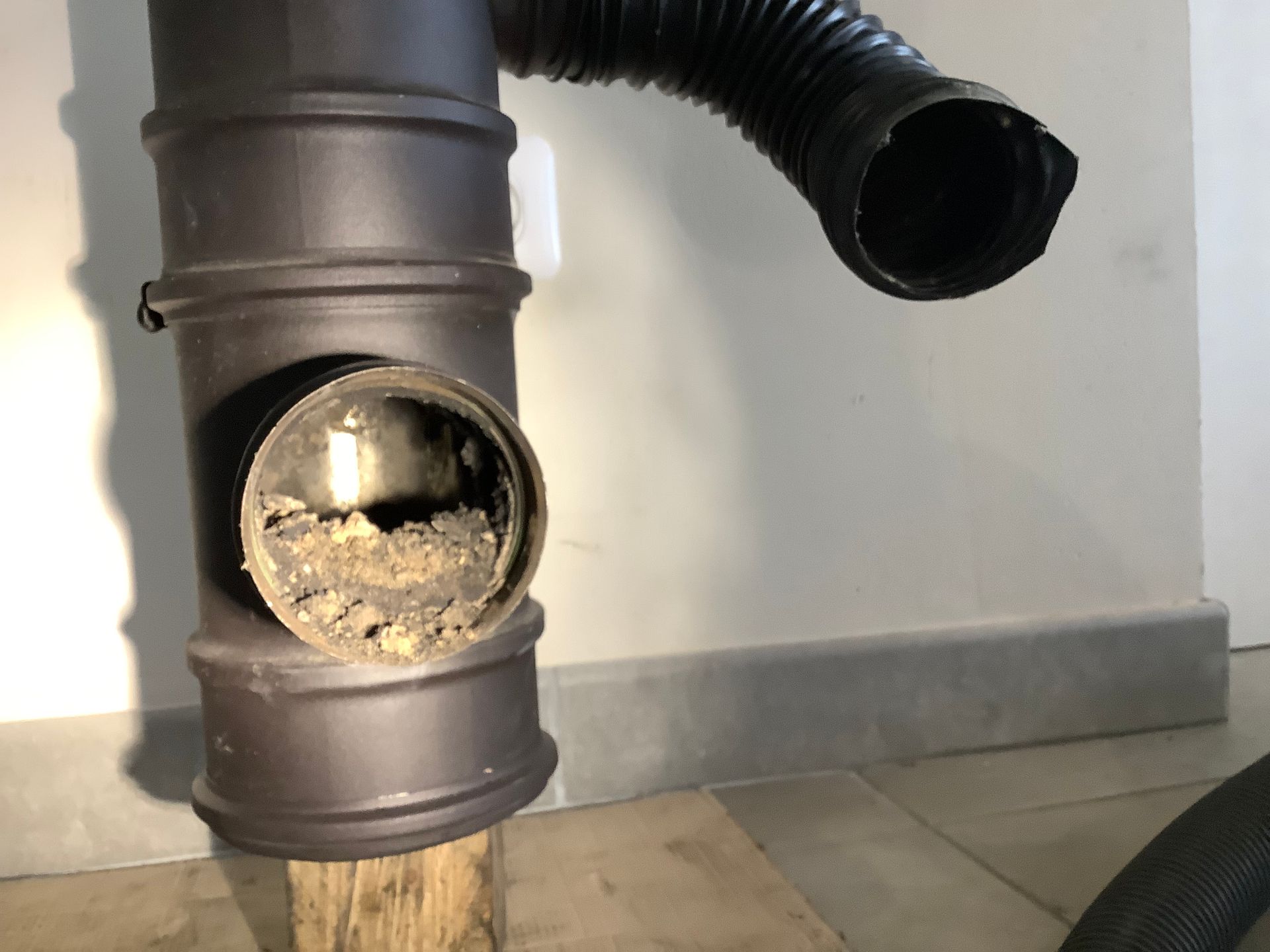 Un raccord en T pour tuyau de poêle en métal foncé, avec un orifice ouvert rempli de débris et un tuyau d'évacuation noir flexible détaché à proximité.