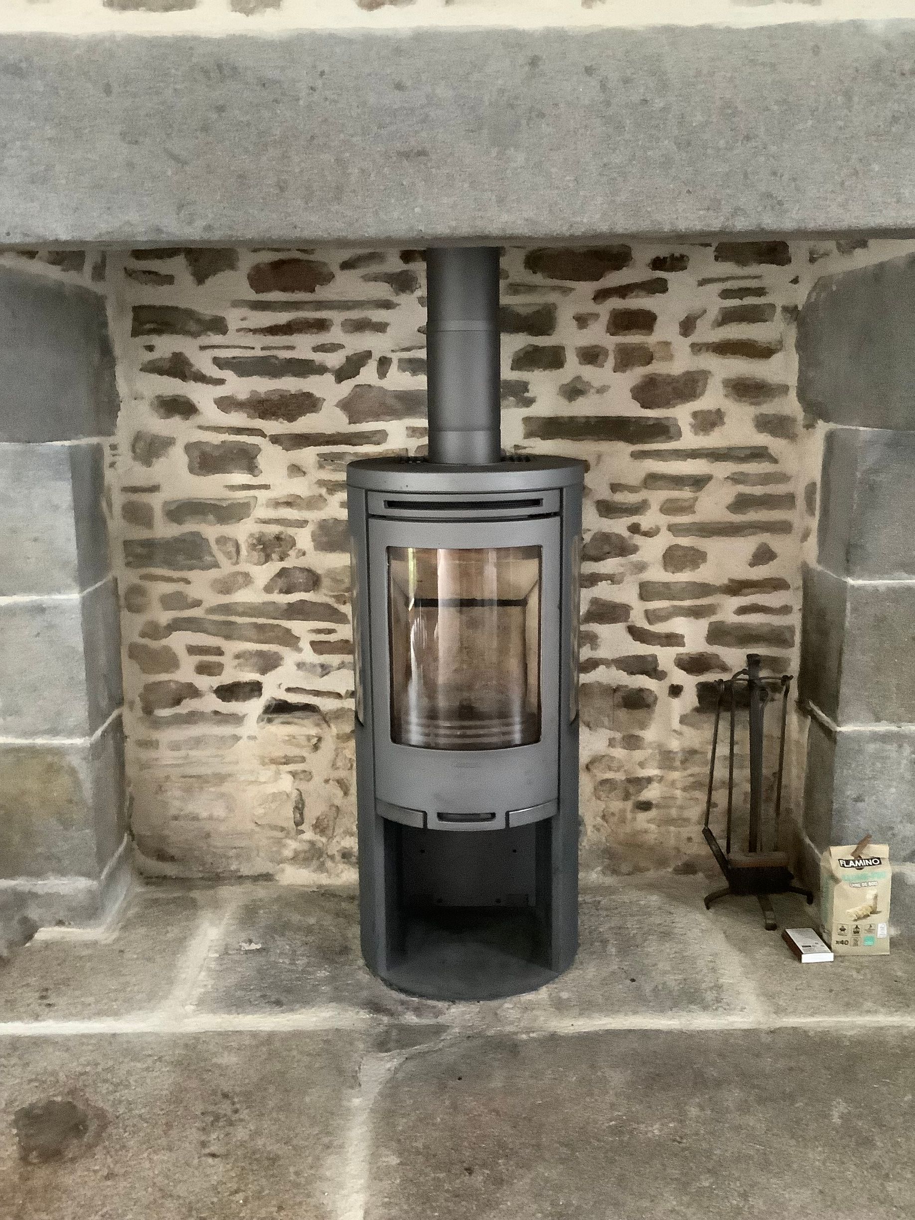 Un poêle à bois cylindrique gris anthracite est placé au centre d'un foyer en pierre.