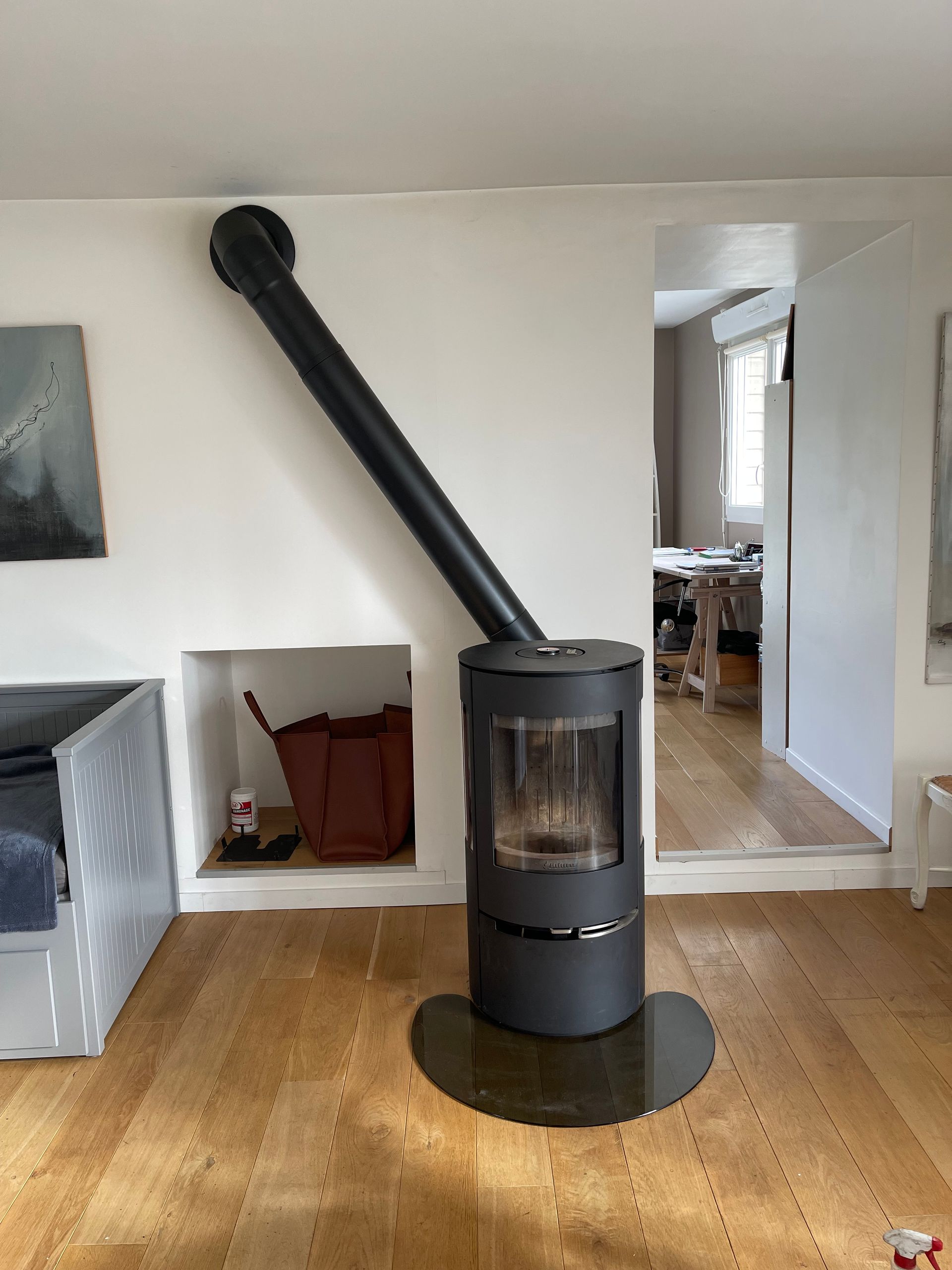 Un poêle à bois cylindrique noir moderne, posé sur une plaque de sol en verre, avec un conduit de fumée s'étendant en diagonale dans le mur.