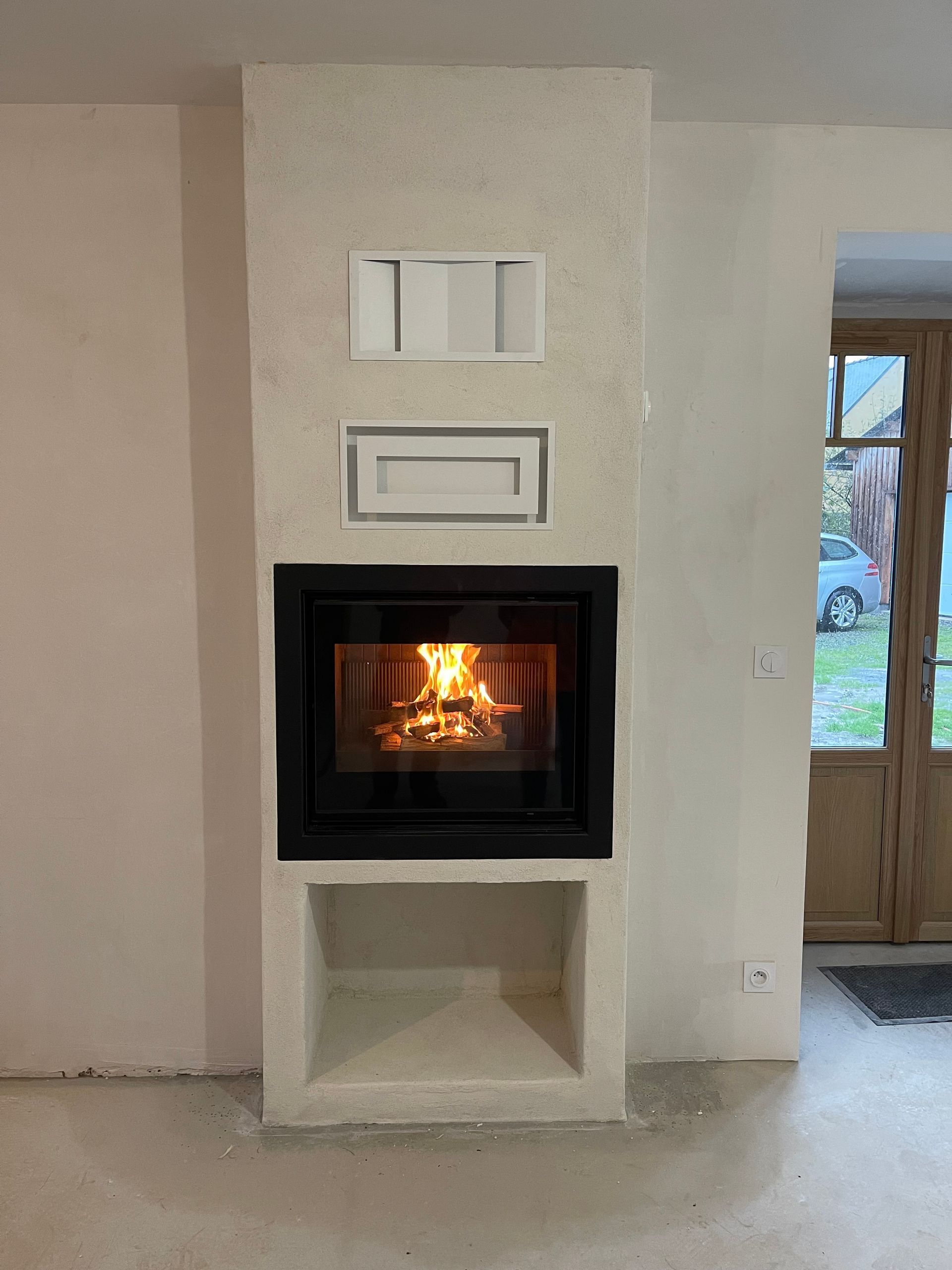 Une cheminée en maçonnerie blanche avec un insert, une niche de rangement pour le bois ouverte en dessous et deux bouches d'aération au-dessus.