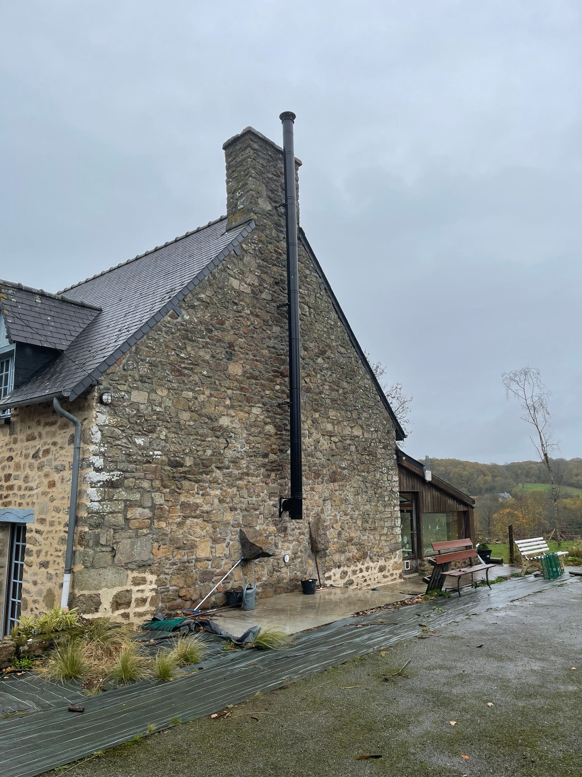 Un mur extérieur en pierre d'une maison, contre lequel est adossée une longue cheminée verticale noire.
