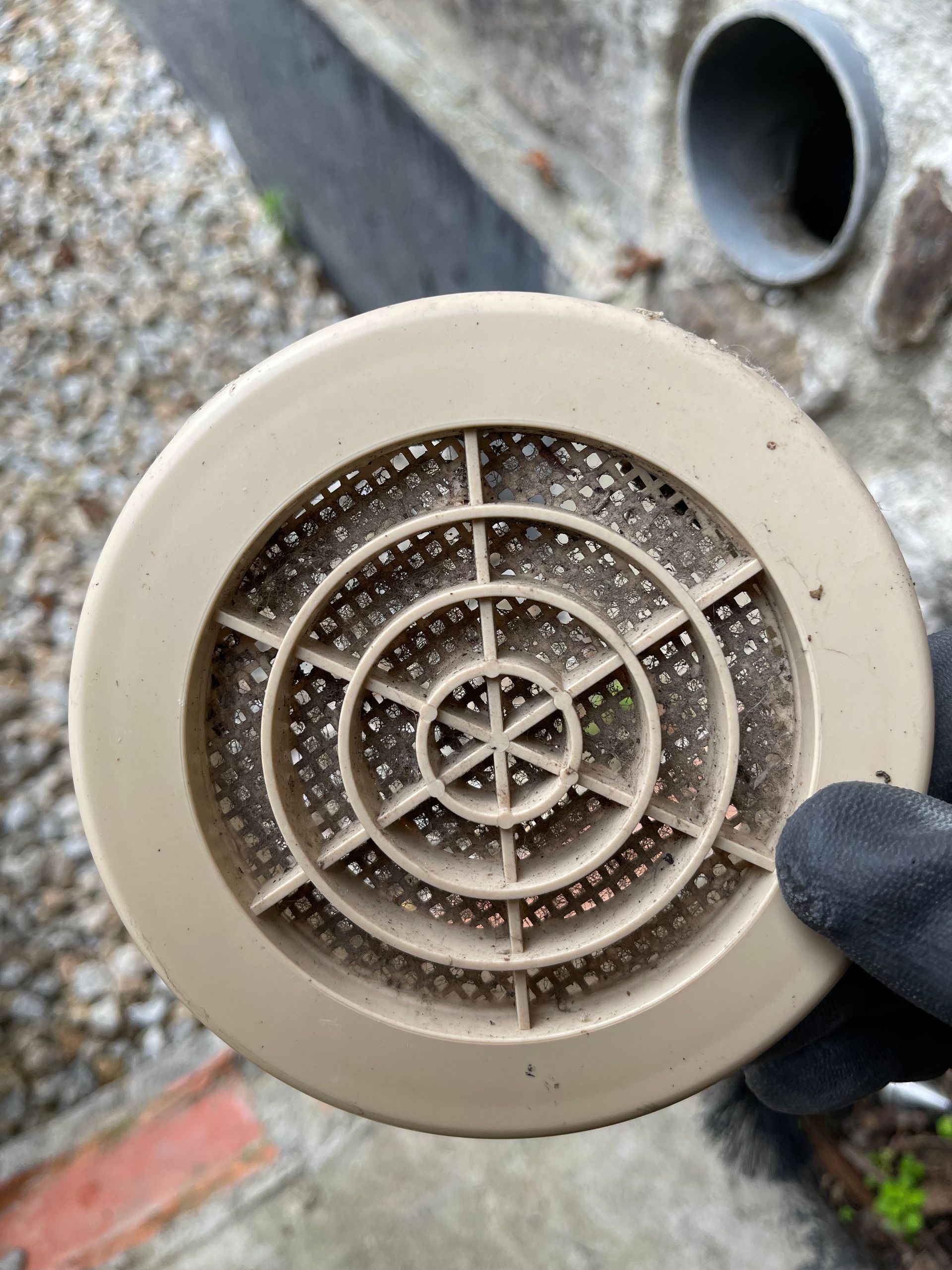 Une main gantée tient une grille d'aération ronde en plastique beige, posée sur un sol de gravier.