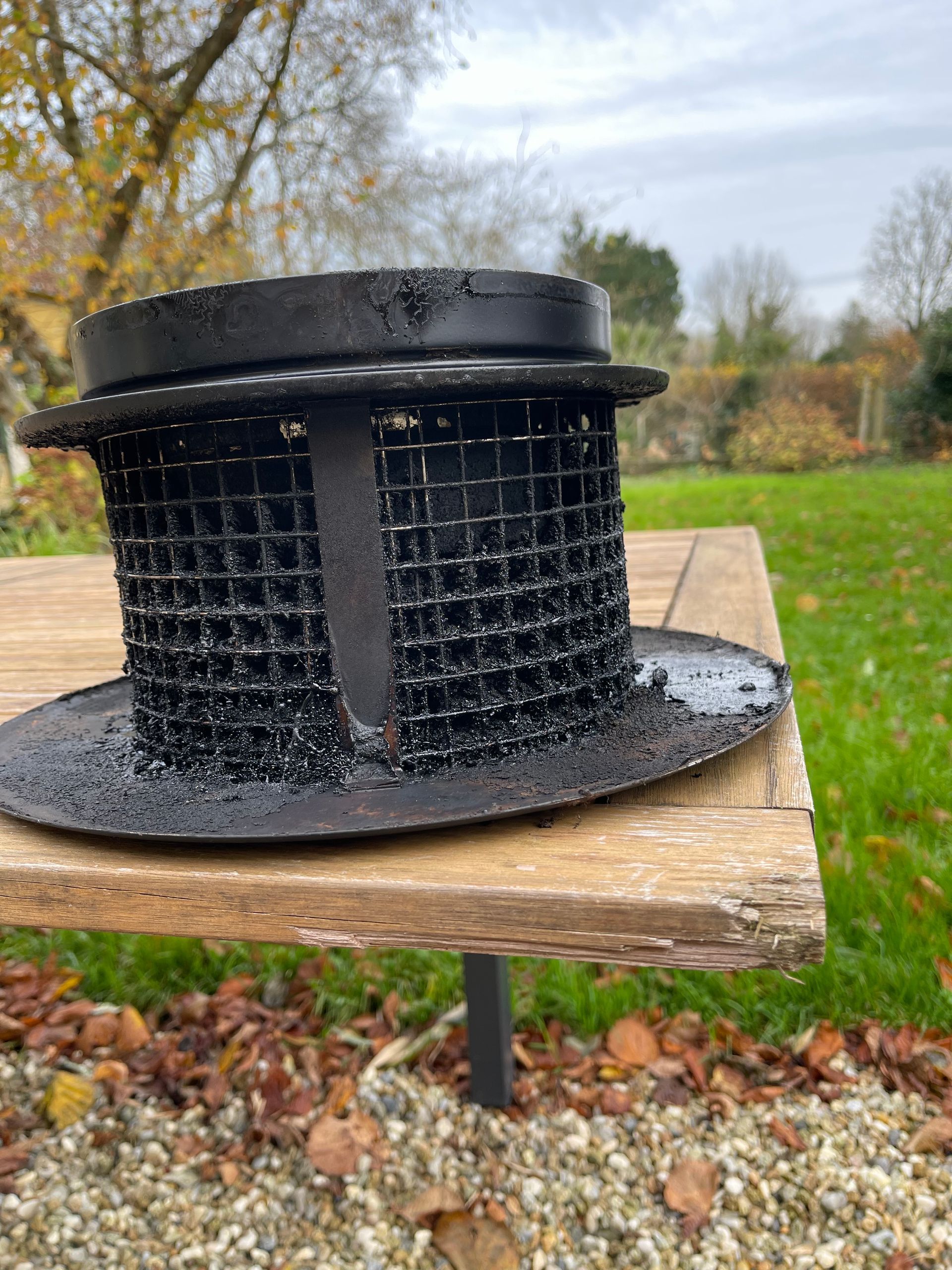 Une cheminée en métal noir avec une grille métallique est posée sur une table de jardin en bois.
