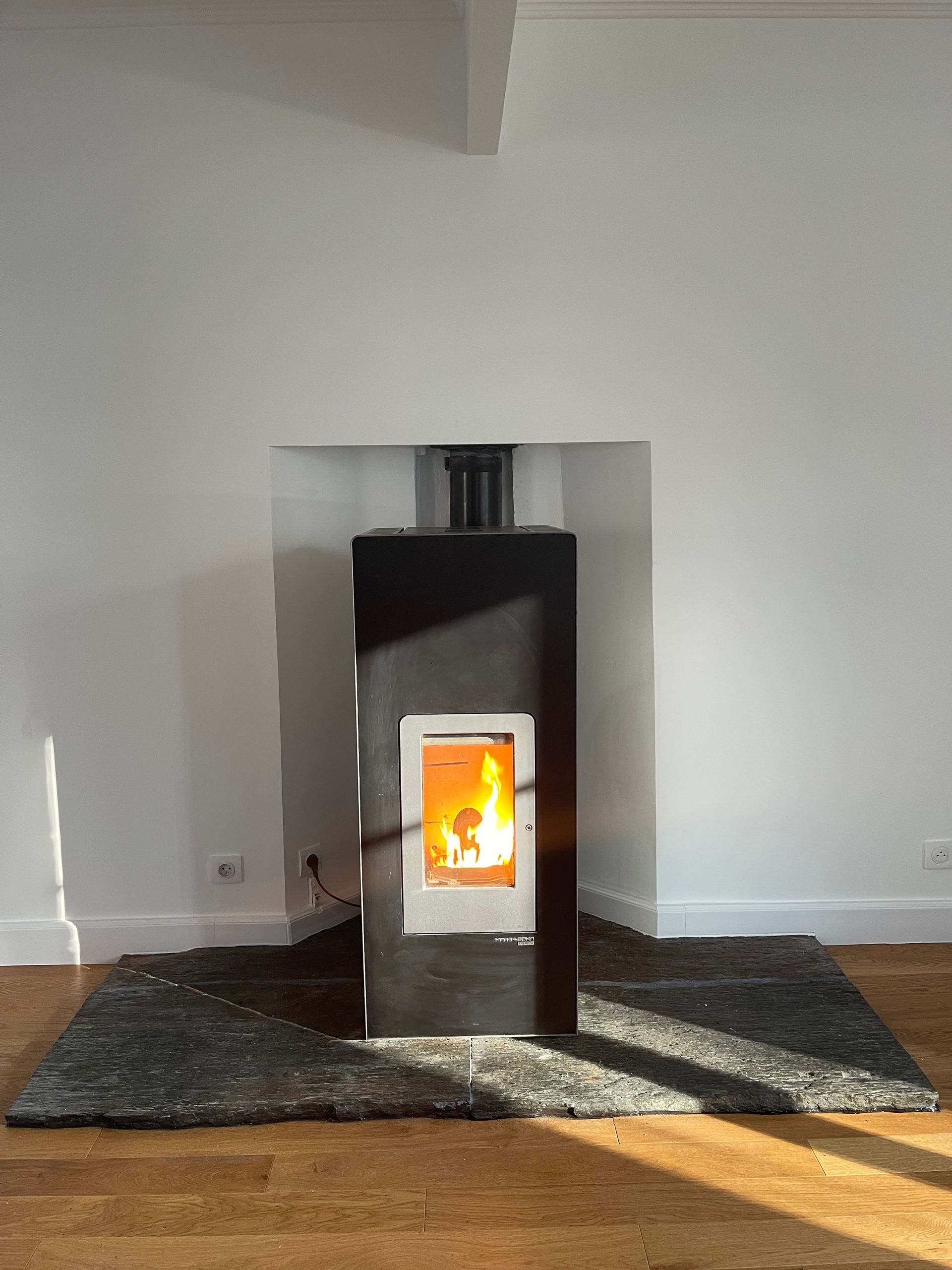 Un poêle à bois noir moderne, avec un feu crépitant, est posé sur un foyer en pierre contre un mur blanc.