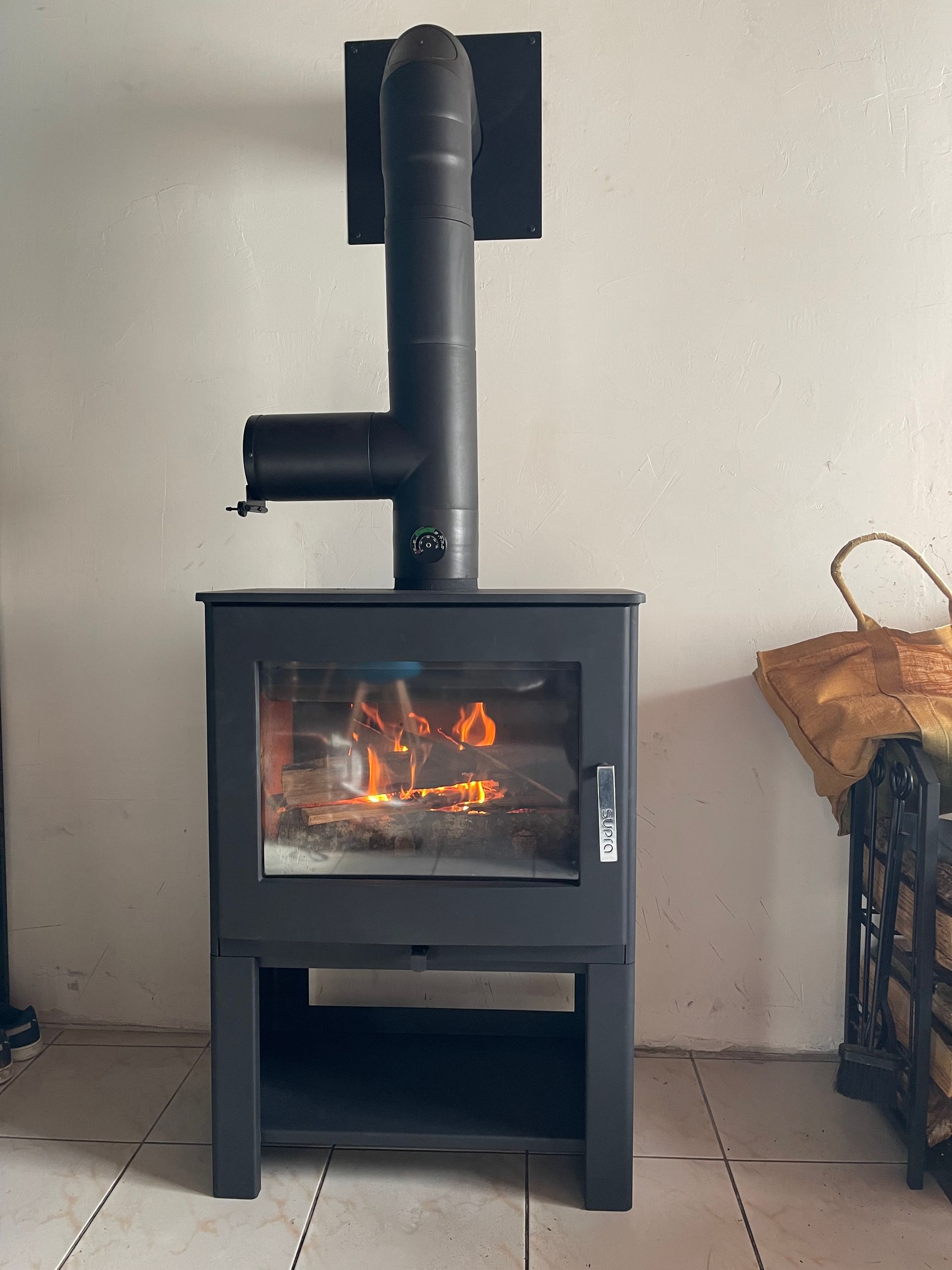 Un poêle à bois noir avec une porte vitrée, d'où s'échappent des flammes chaudes, est placé contre un mur blanc avec une sortie de cheminée.