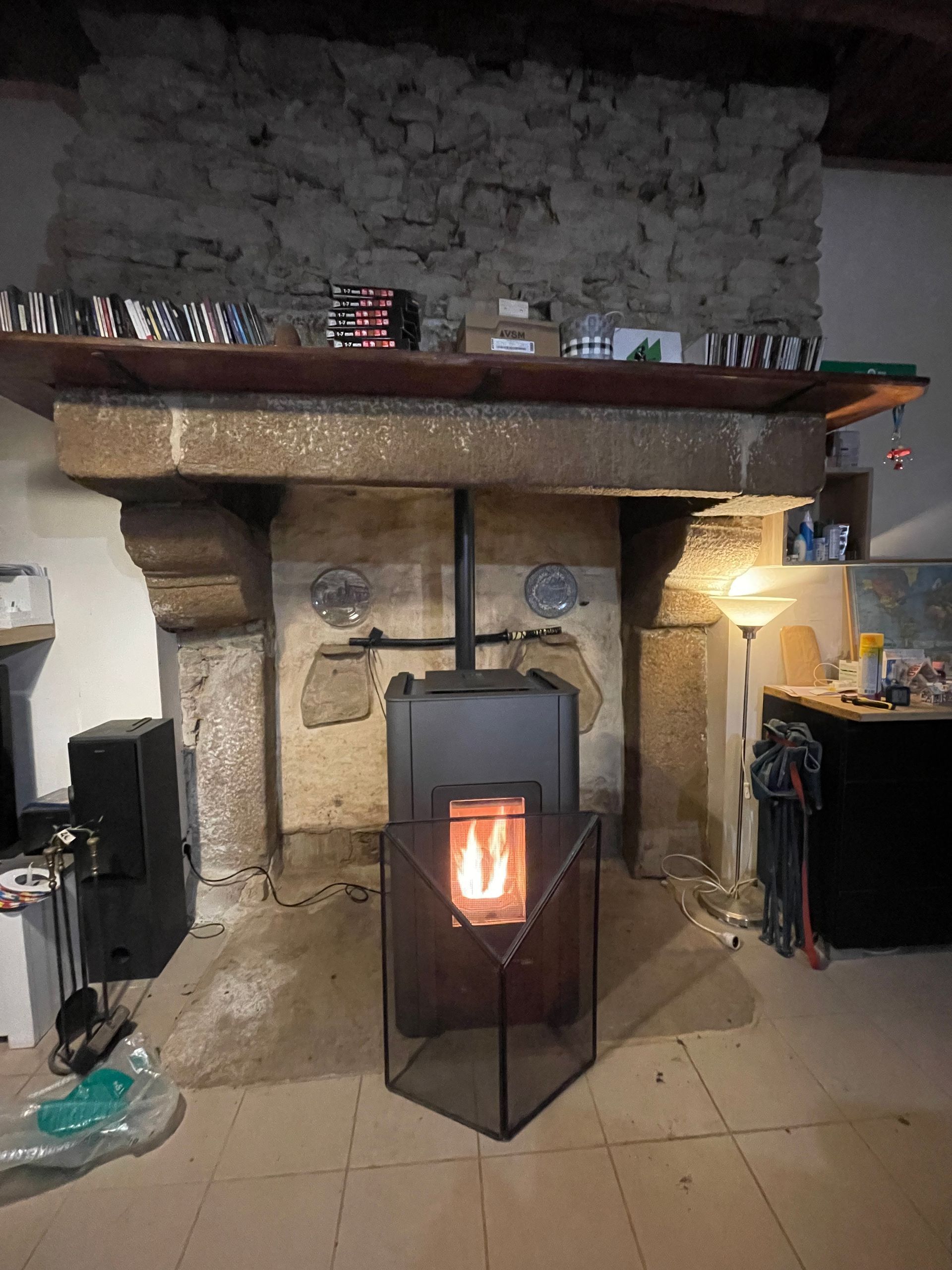 Un poêle à granulés, dont la flamme est vive, se dresse à l'intérieur d'une grande cheminée rustique en pierre.