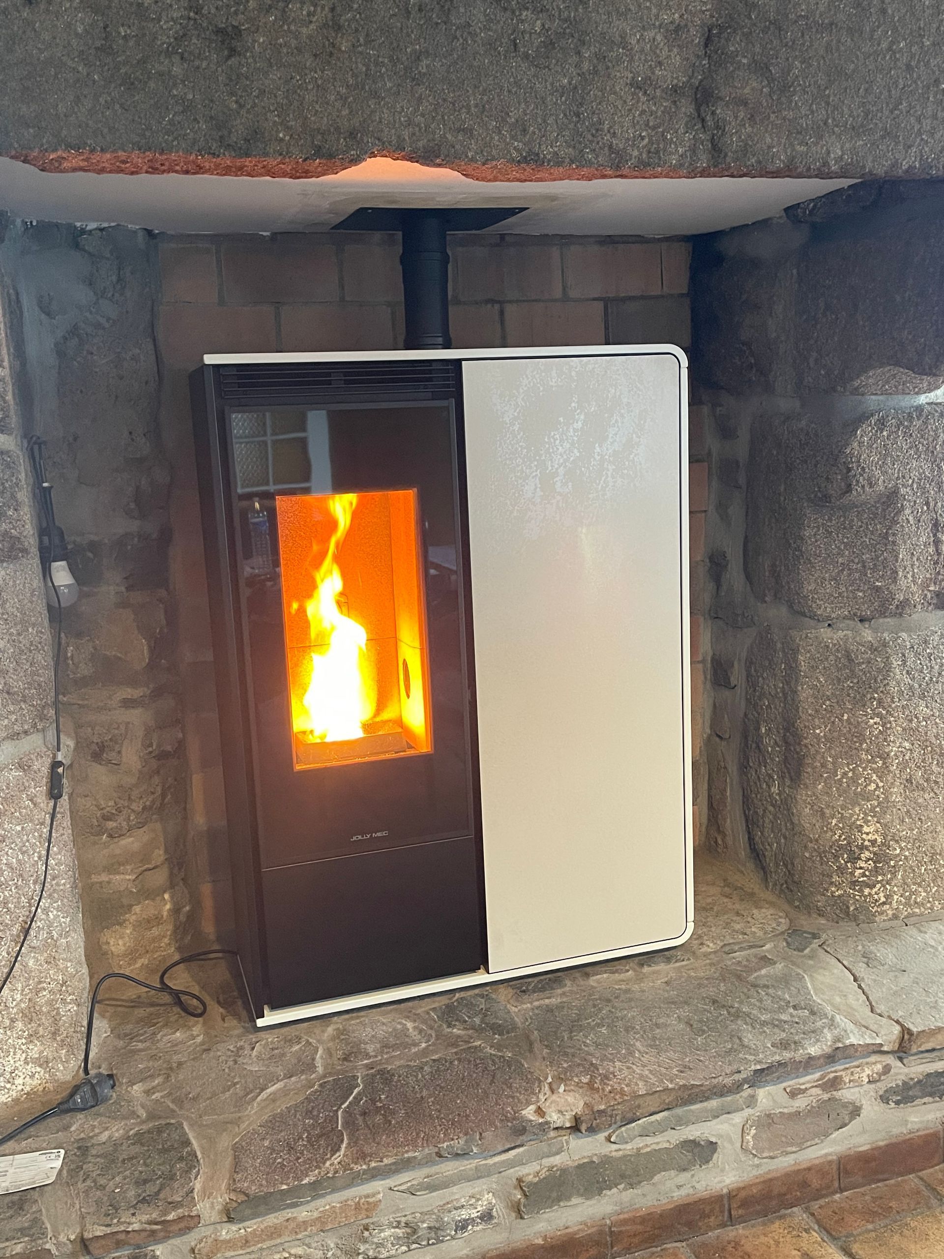 Un poêle à granulés moderne, avec une porte en verre noir et un panneau latéral blanc, brûle intensément dans une cheminée rustique en pierre.