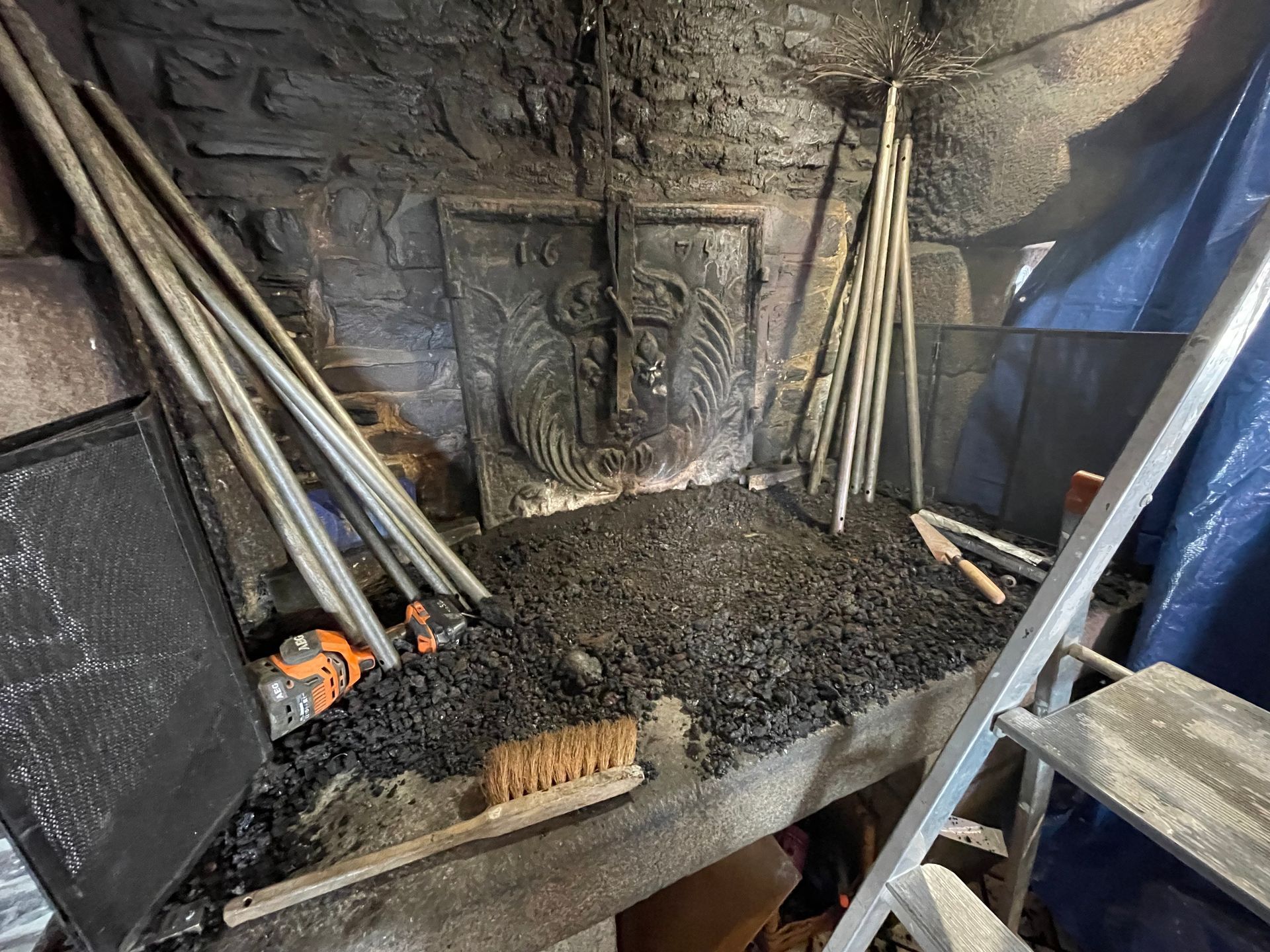 Une grande cheminée avec une plaque de fond décorative en fonte, remplie de charbon de bois épars, de cendres, d'outils et d'une brosse de nettoyage.