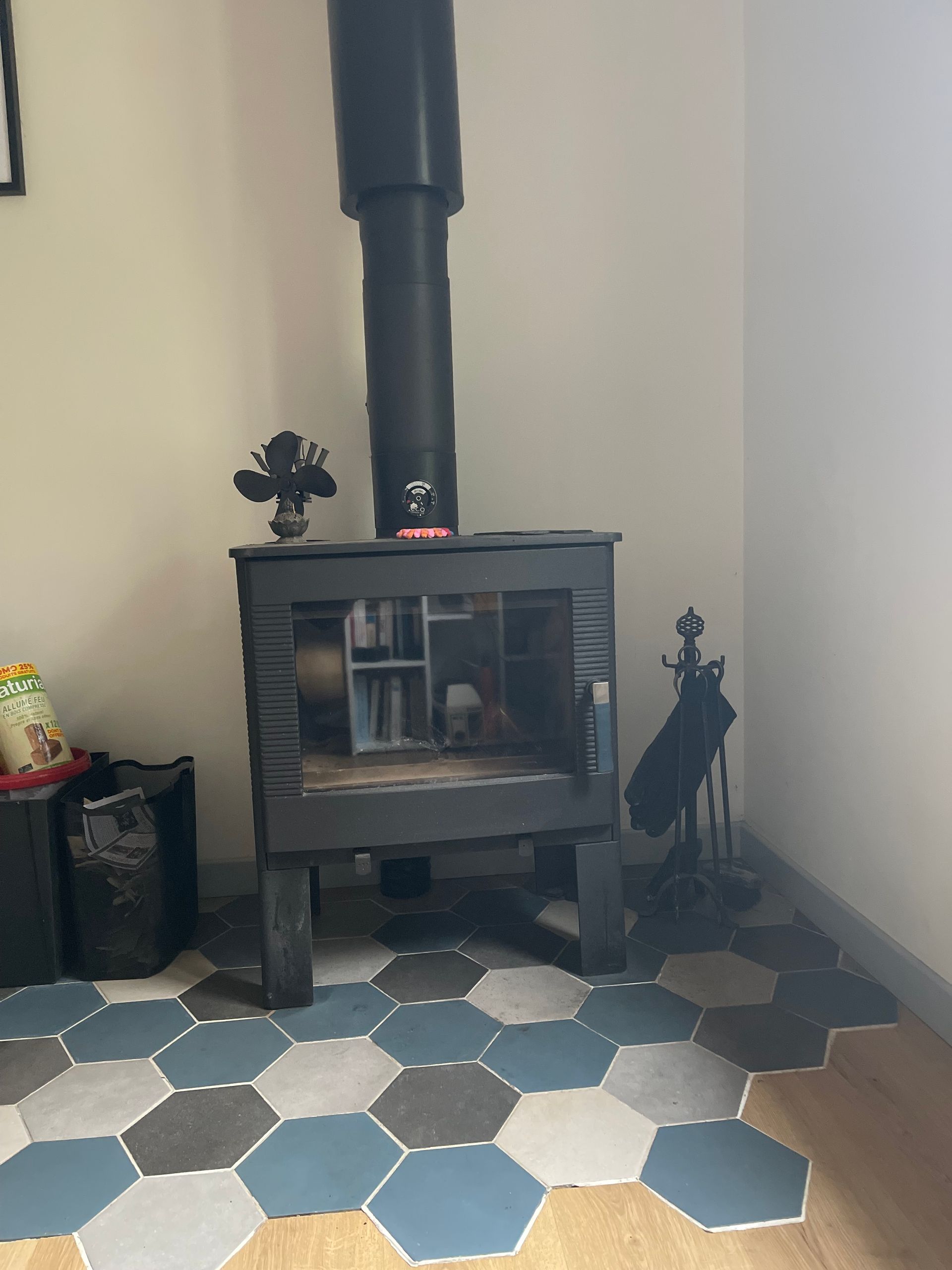 Un poêle à bois noir est installé sur un foyer en carreaux hexagonaux bleus, gris et blancs, dans un coin de la pièce.