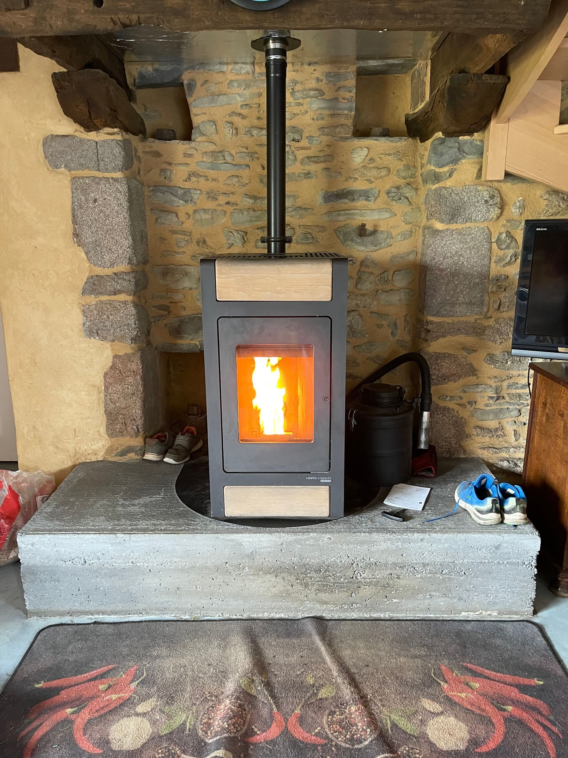 Un poêle à granulés allumé, aux panneaux beiges, est installé sur un foyer en pierre à l'intérieur d'une cheminée rustique aux murs en pierre apparente.