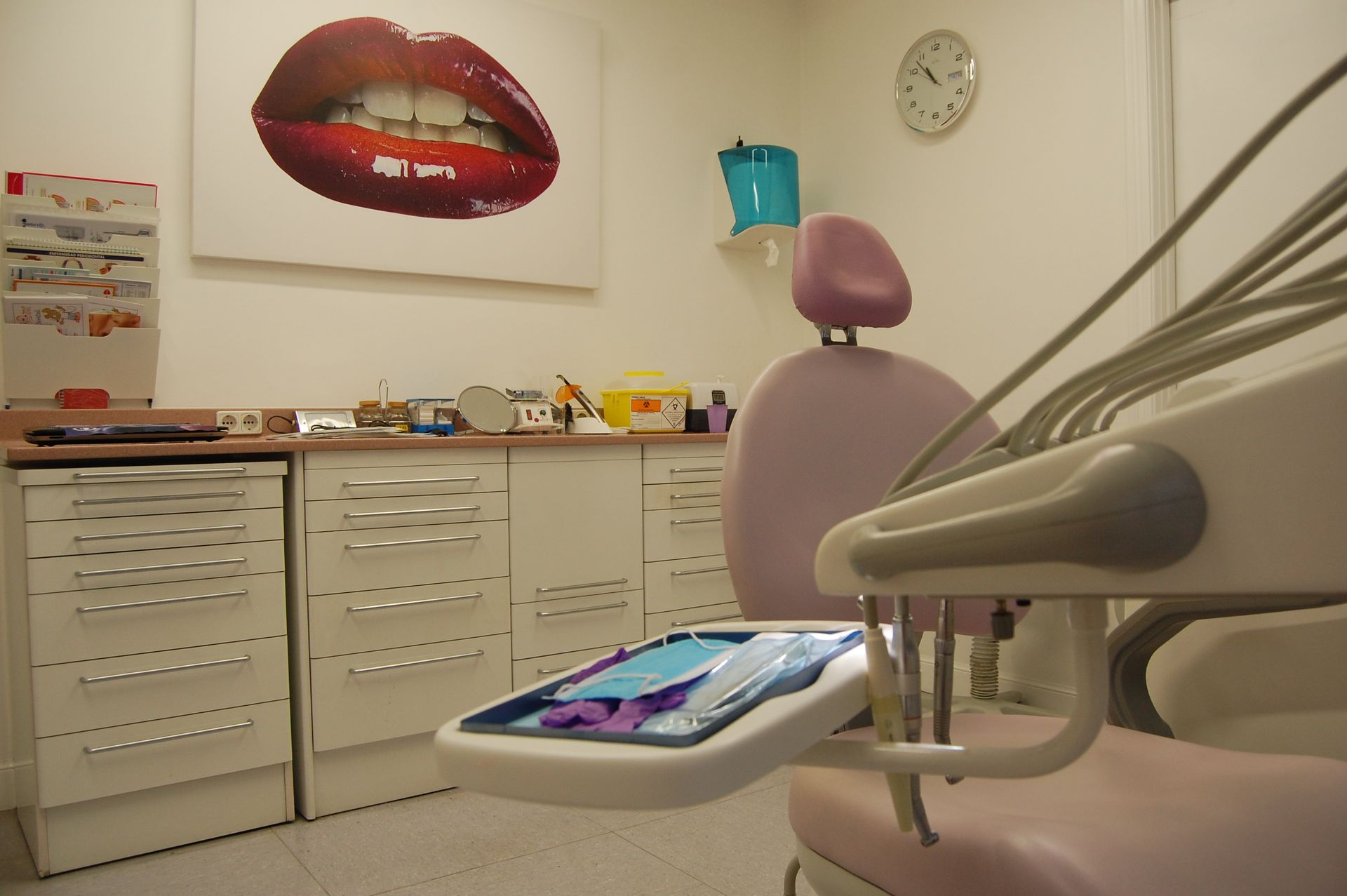 Sala de examen dental vacía con una silla rosa, gabinetes blancos y un gran póster de labios en la pared.