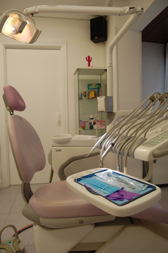 Sala de exploración dental con sillón rosa, luz de techo y bandeja de instrumentos cerca del lavabo.
