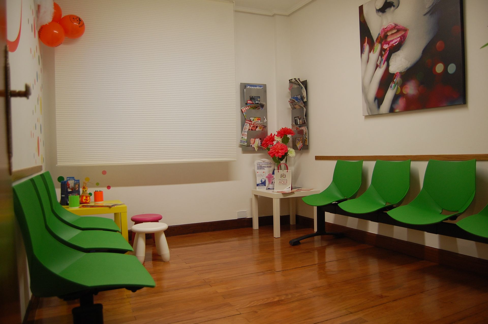Sala de espera con sillas verdes, suelo de madera, mesa pequeña y coloridos cuadros en la pared.