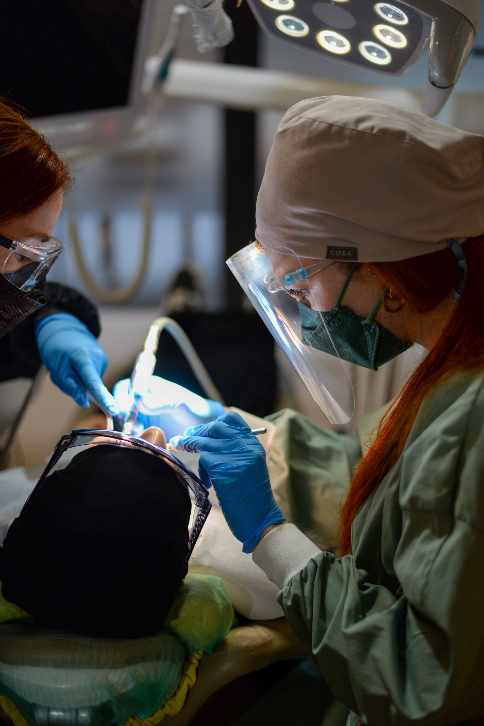 Dentista y asistente trabajando en los dientes de un paciente bajo una luz brillante de examen, usando mascarillas y guantes.