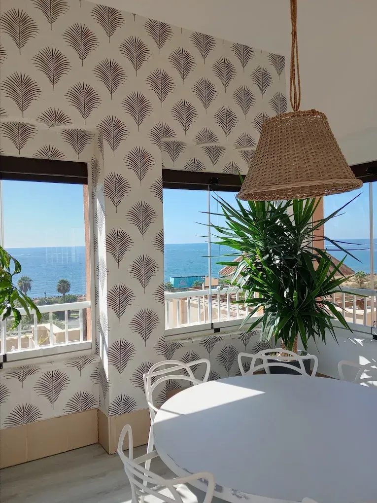 Luminoso rincón comedor junto al mar con mesa blanca, lámpara colgante de mimbre y vistas al océano a través de las ventanas.