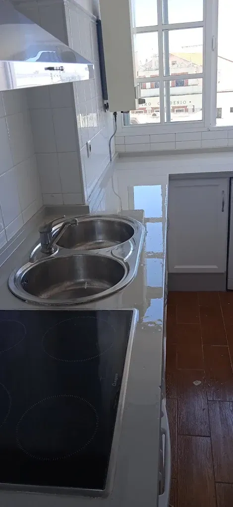 Cocina pequeña con fregadero de acero inoxidable, armarios blancos, suelo de baldosas y una ventana al fondo.