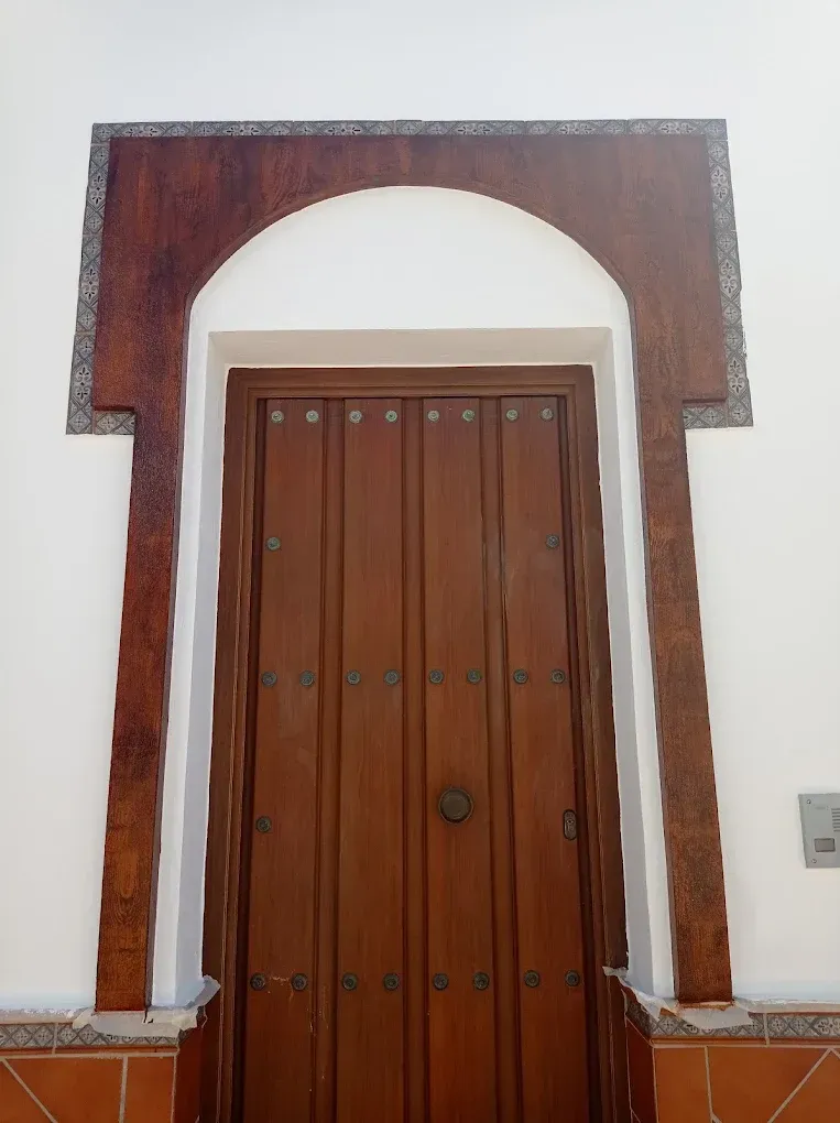 Entrada arqueada de madera con puerta de paneles marrones enmarcada en una pared blanca.
