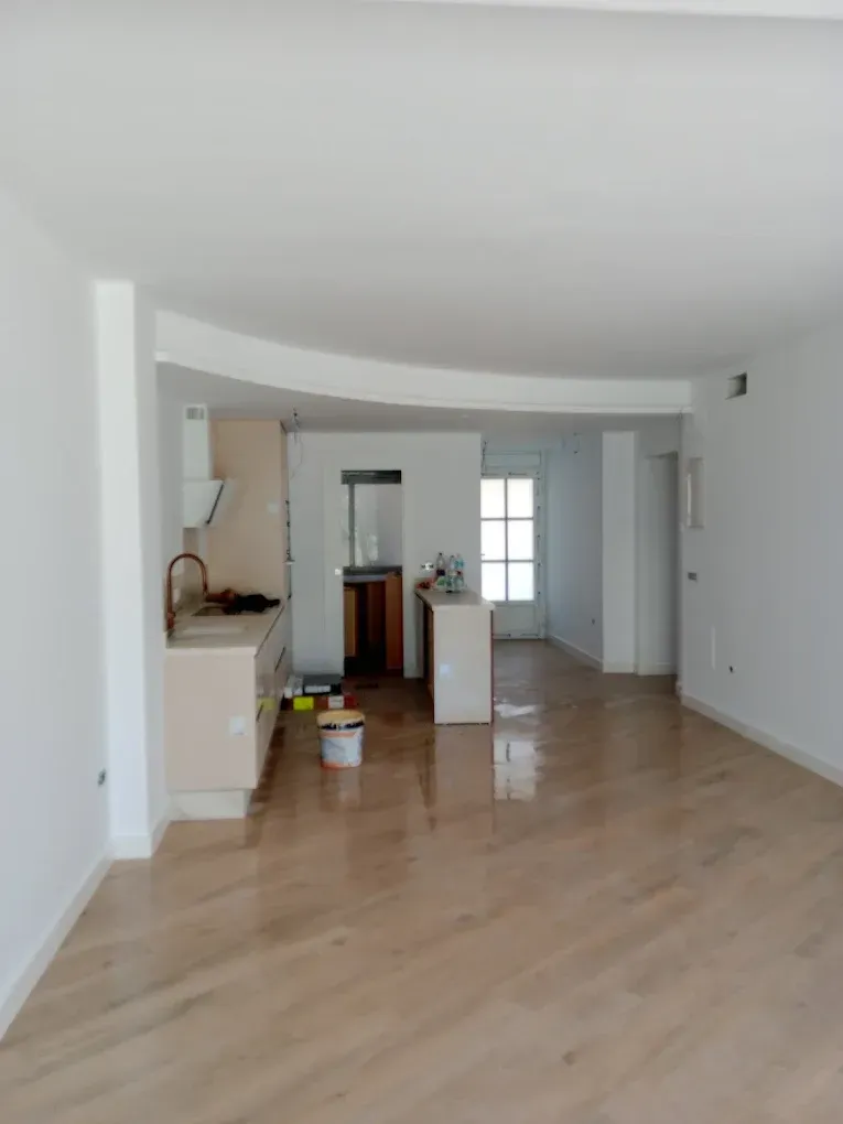 Sala de estar blanca y vacía con pisos de madera clara y una pequeña área de cocina en la parte trasera.