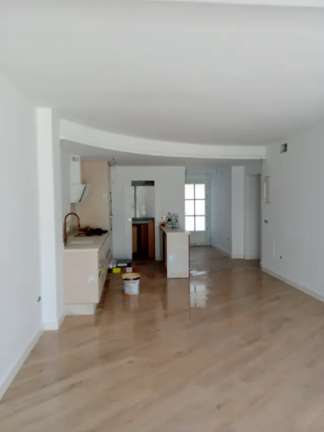 Sala de estar blanca y vacía con pisos de madera clara y una pequeña área de cocina en la parte trasera.