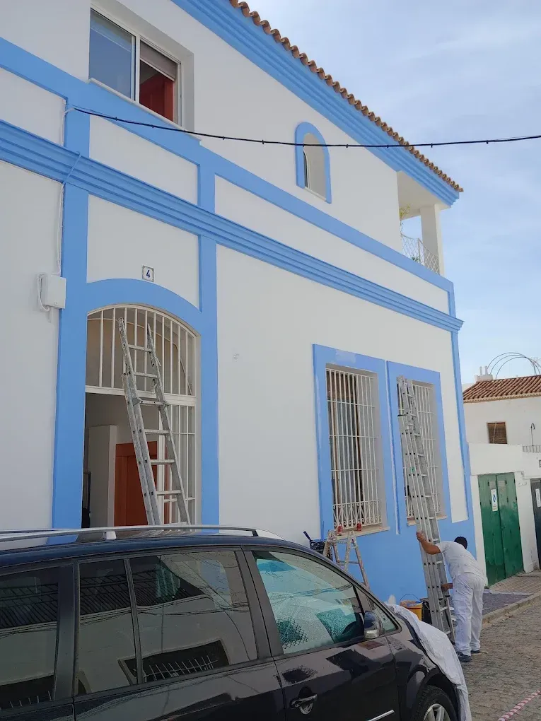 Fachada de una casa azul y blanca con ventanas arqueadas, un coche aparcado y una persona de pie junto a la entrada.