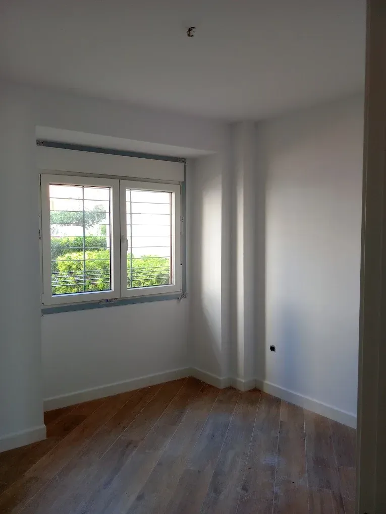 Habitación blanca vacía con suelo de madera y una ventana con vistas a la vegetación.