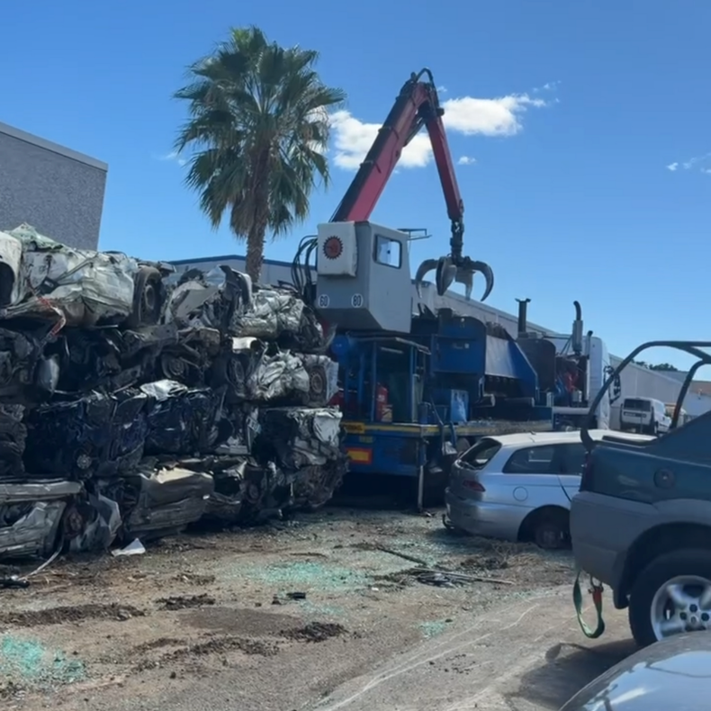 Une casse automobile avec une grue soulevant des voitures broyées à côté d'un palmier, sous un ciel bleu.