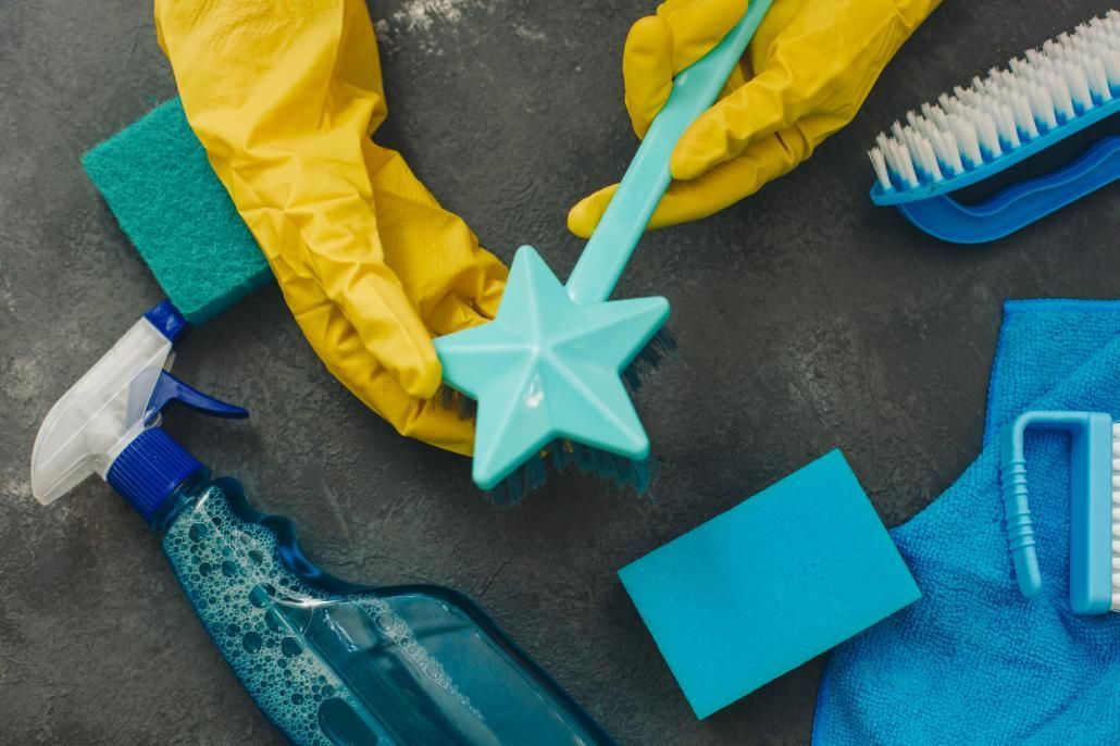 Unas manos con guantes amarillos sostienen un cepillo de limpieza entre otros productos de limpieza de color azul y verde.