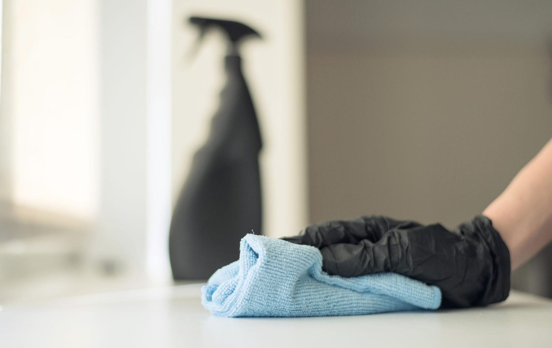 Persona con guantes negros limpiando una superficie blanca con un paño azul; botella de spray en el fondo.