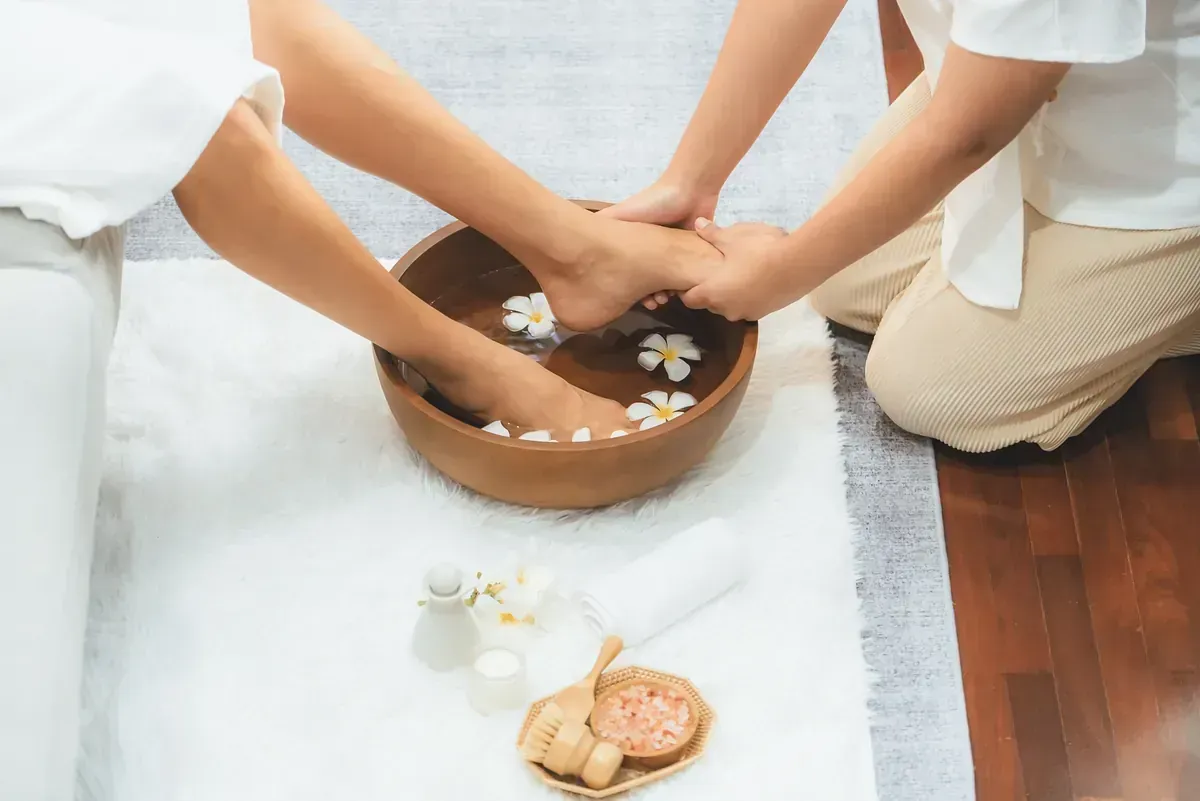 Persona recibiendo masaje de pies en un recipiente de madera con agua y flores; entorno de spa.