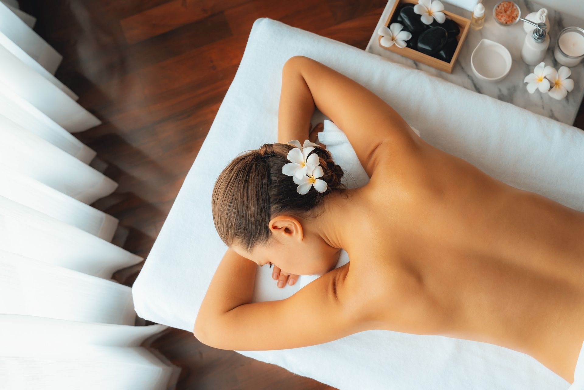 Mujer recibiendo tratamiento de spa; acostada boca abajo, flor en el cabello, sábana blanca, mesa de madera con tratamientos.