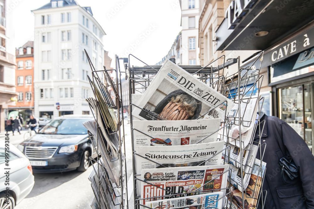 Zeitungskiosk auf einer europäischen Straße mit deutschen Zeitungen, Gebäuden und einem Auto.