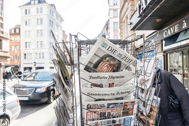 Zeitungskiosk auf einer europäischen Straße mit deutschen Zeitungen, Gebäuden und einem Auto.