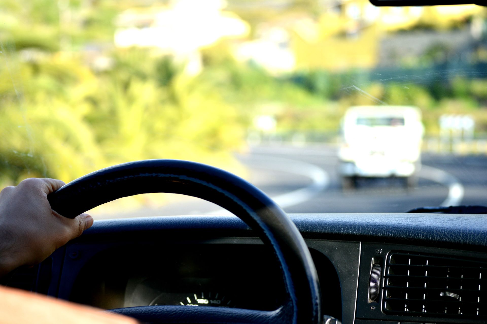 Une main agrippée à un volant noir, roule sur une route sinueuse. Un autre véhicule est devant moi sur le chemin verdoyant et ensoleillé.