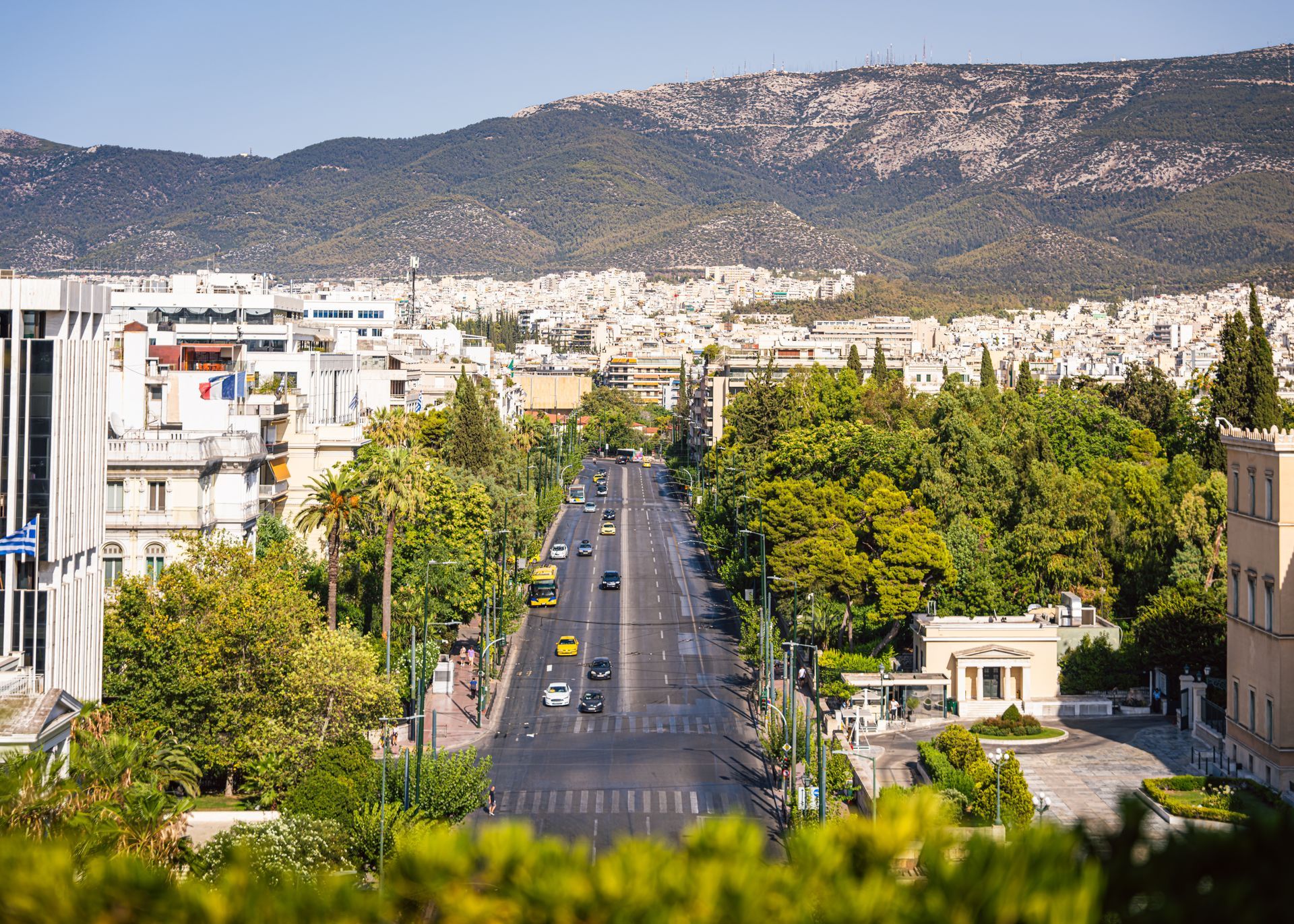 Vue aérienne d'une longue avenue, avec des voitures et des bâtiments, des arbres verts et une montagne en arrière-plan.