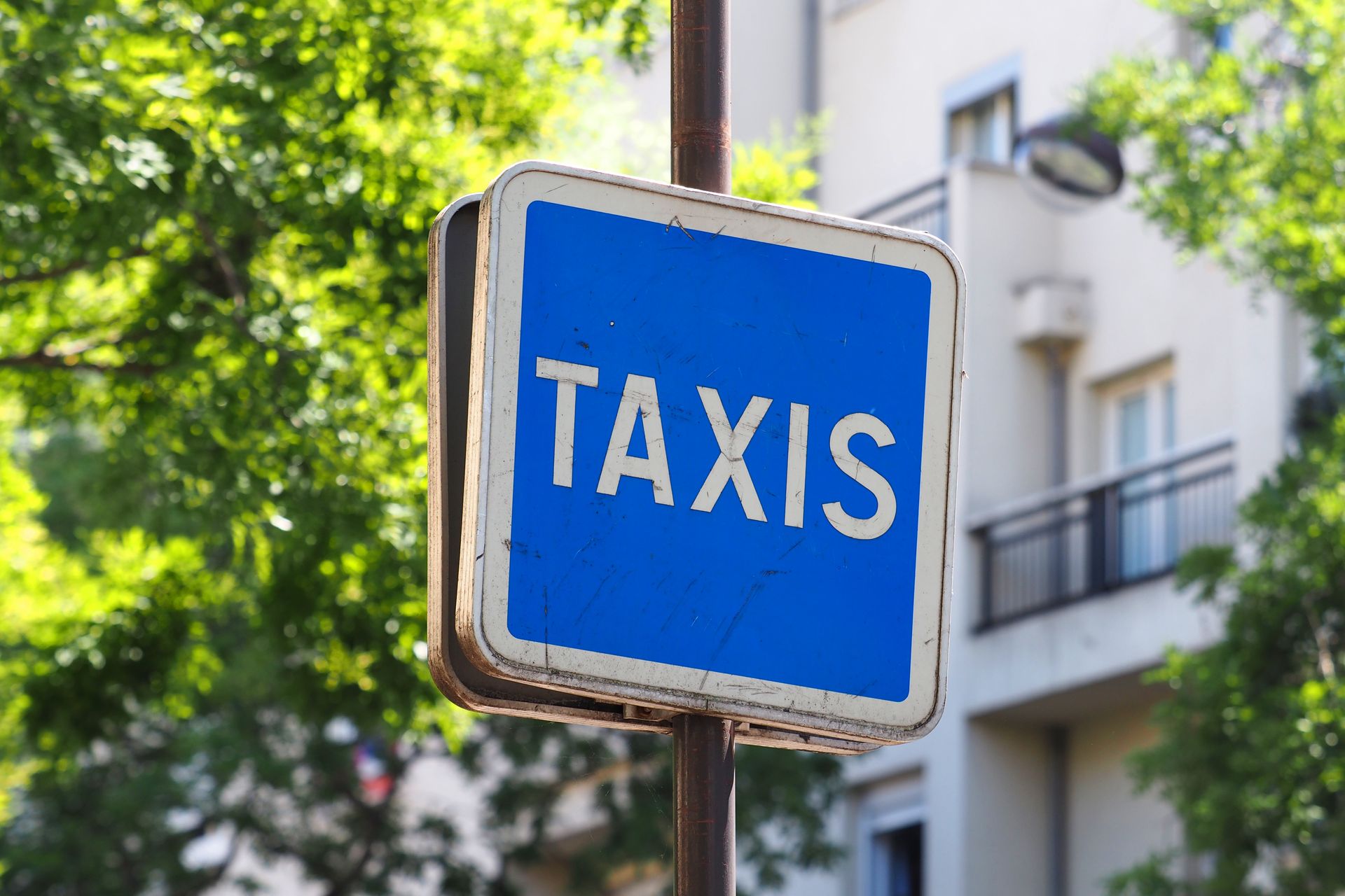 Panneau de taxi bleu et blanc sur un poteau ; devant un bâtiment et des arbres.