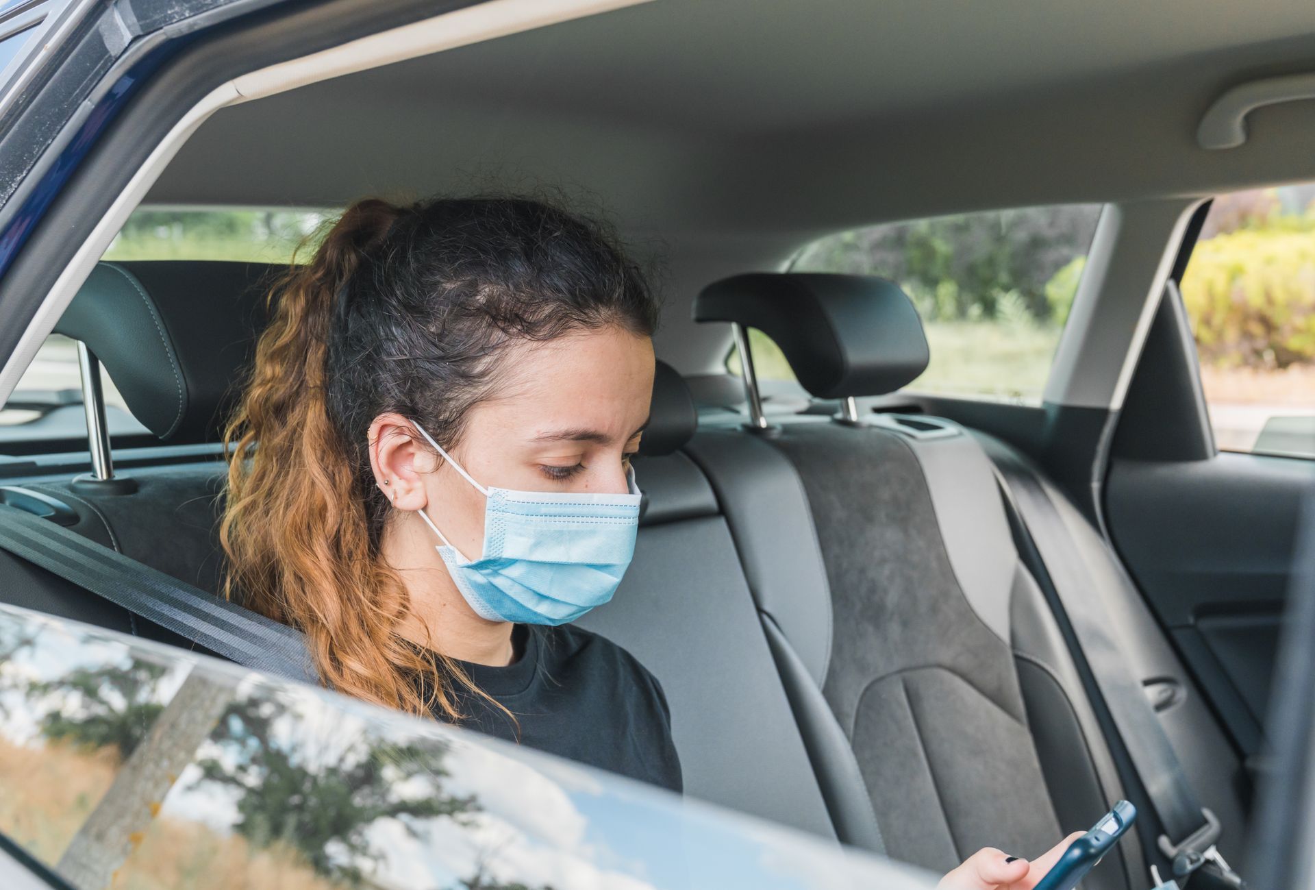 Une femme portant un masque sur la banquette arrière d'une voiture regarde un téléphone.