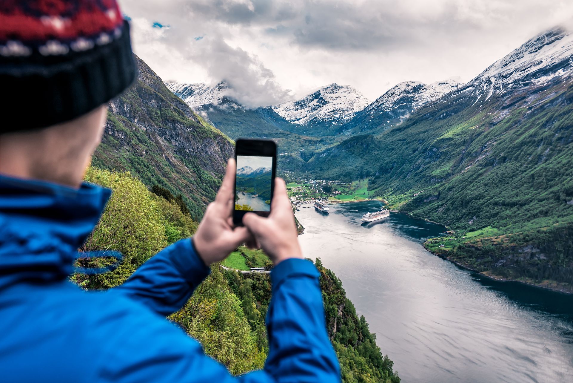Un homme en veste bleue photographie un fjord pittoresque avec son téléphone. On aperçoit des montagnes enneigées, une vallée verdoyante et des bateaux de croisière.