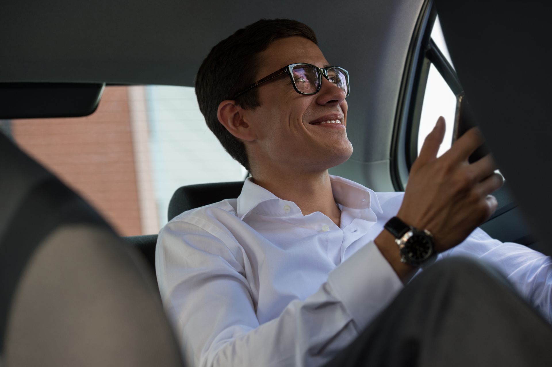 Un homme dans une voiture portant des lunettes, une chemise blanche et une montre, souriant tout en regardant un téléphone.