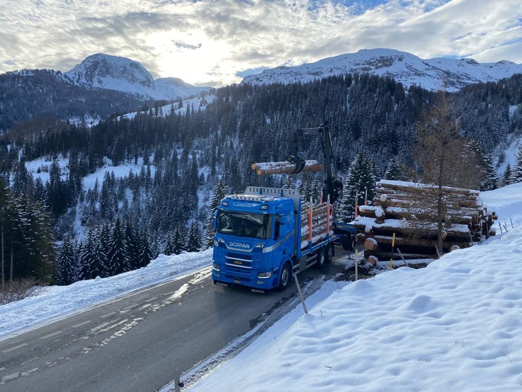 Ein blauer Lastwagen fährt eine verschneite Bergstraße entlang.