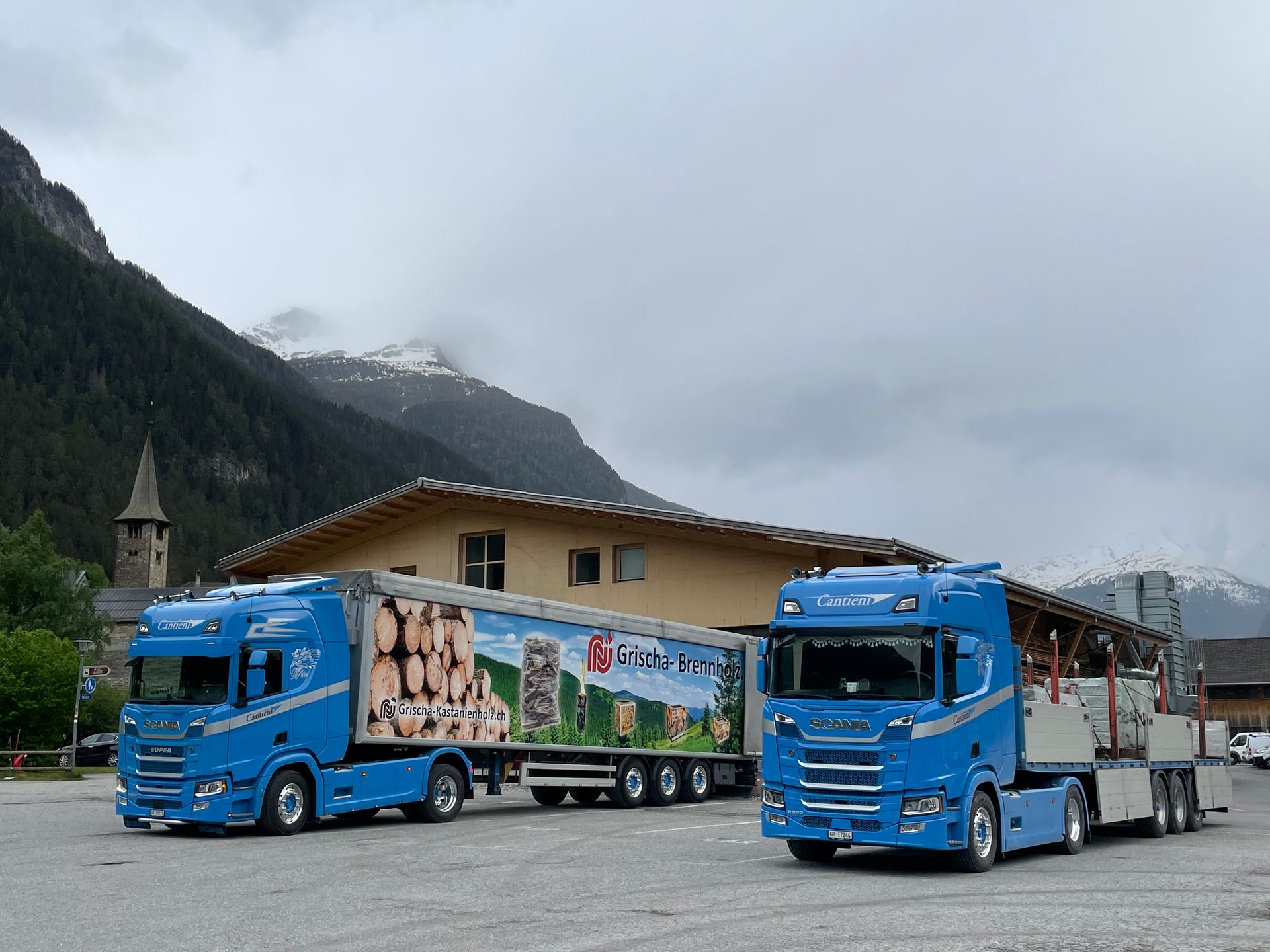 Zwei blaue Lastwagen parken vor einem Gebäude mit Bergen im Hintergrund.