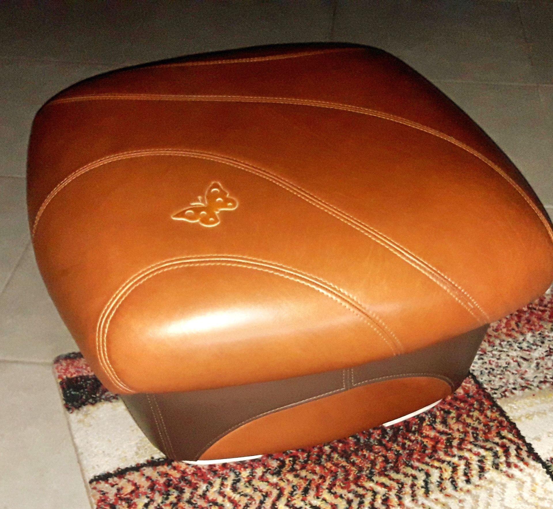 Pouf en cuir marron avec surpiqûres blanches et petit emblème décoratif, posé sur un tapis à motifs.