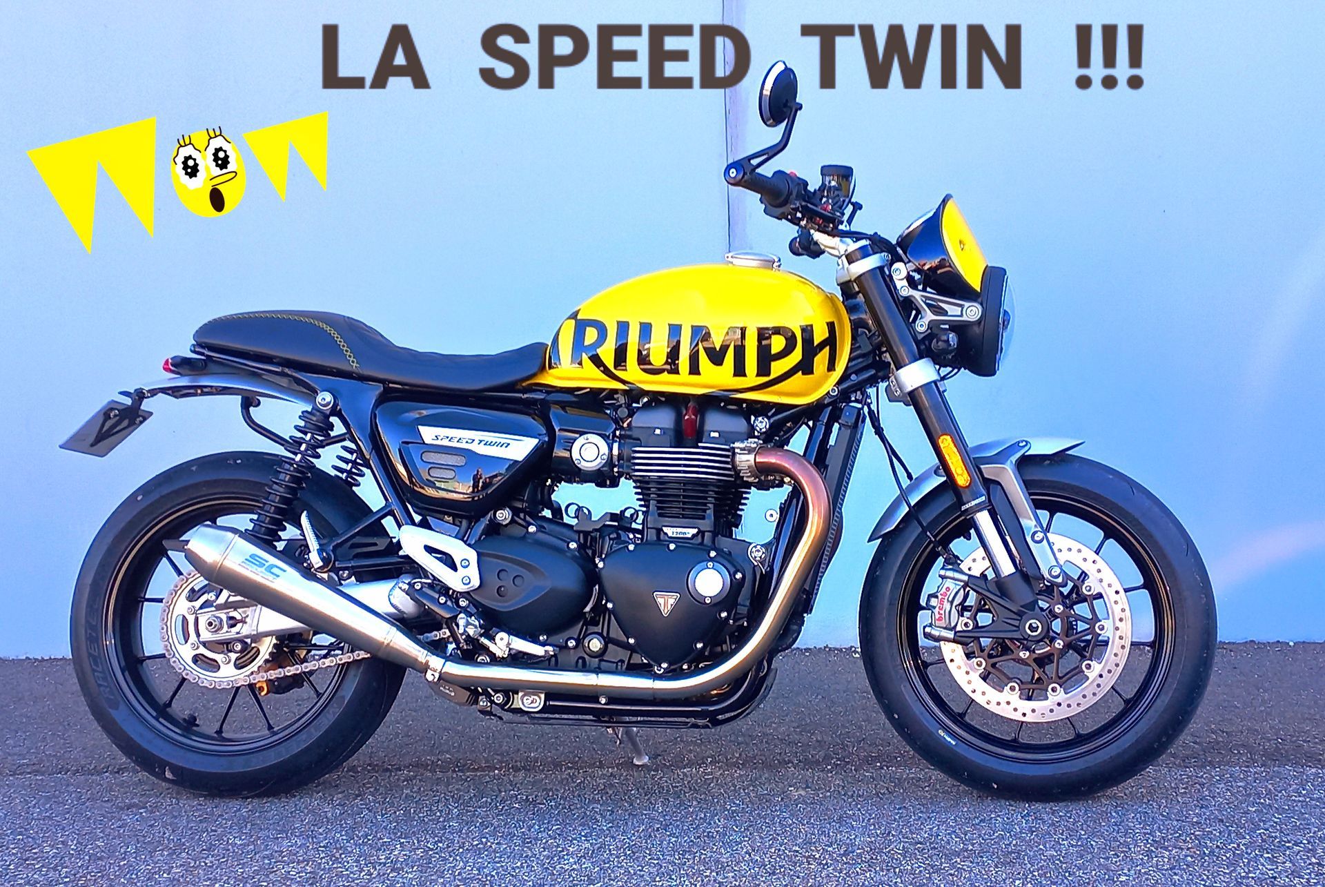 Moto Triumph noire sur fond bleu avec l'inscription LA SPEED TWIN !!! et des drapeaux jaunes