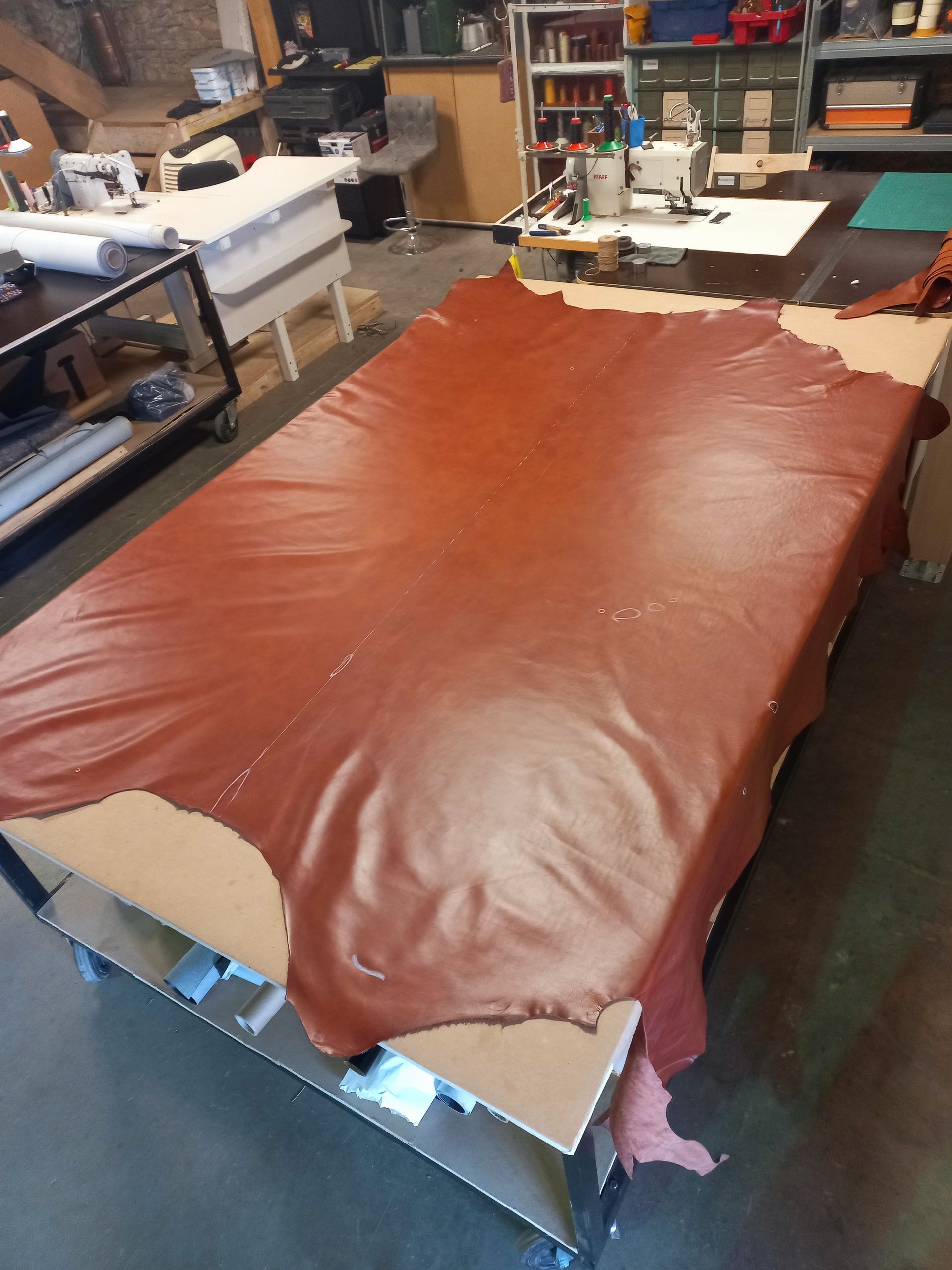 Une grande peau de cuir marron est étendue sur une table de travail dans un atelier, entourée d'outils et de matériaux.