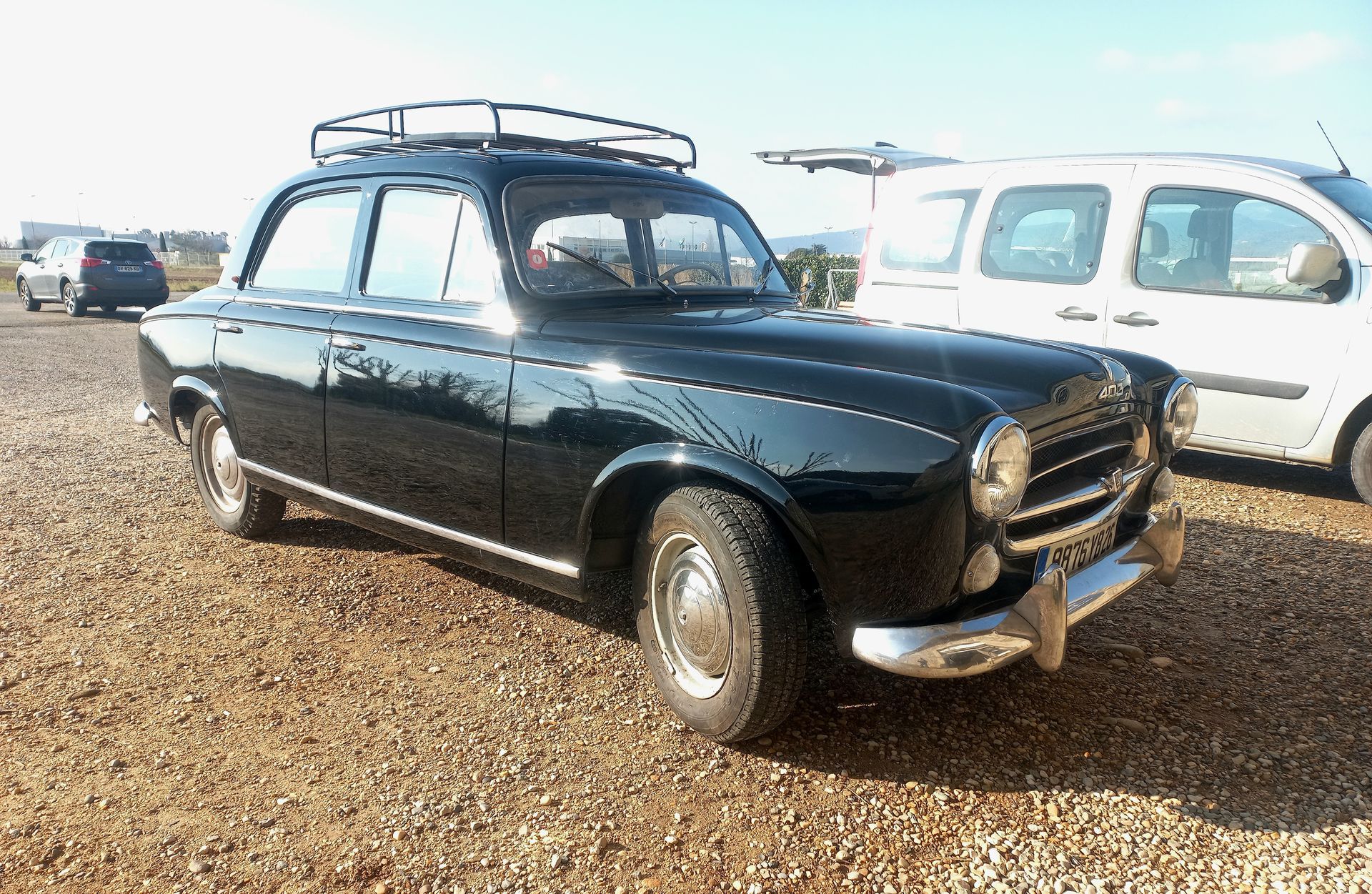 Voiture ancienne noire avec barres de toit garée sur un terrain gravillonné à côté d'une camionnette blanche